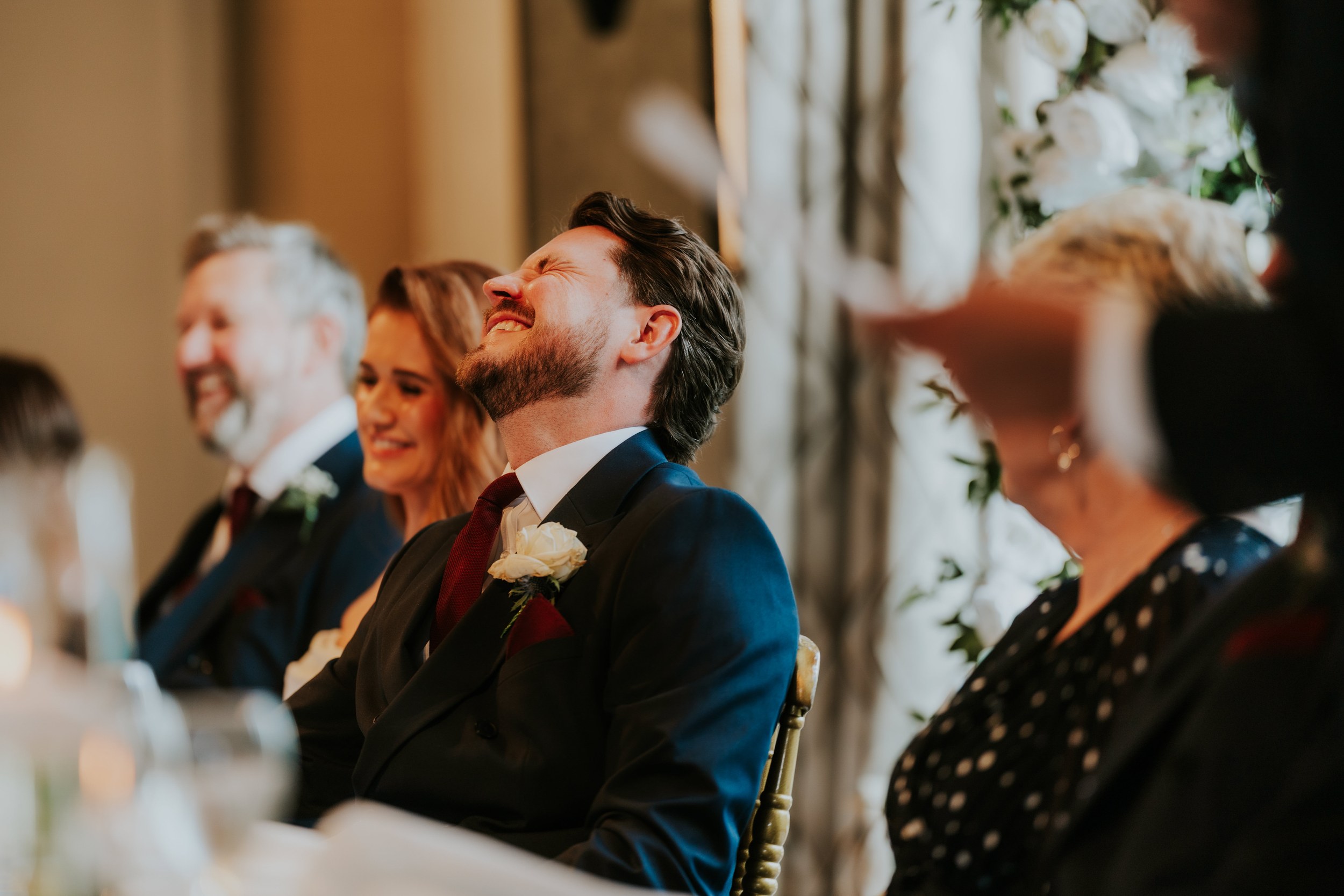 Groom Jack laughing with eyes closed at head table, navy suit and white rose boutonniere.