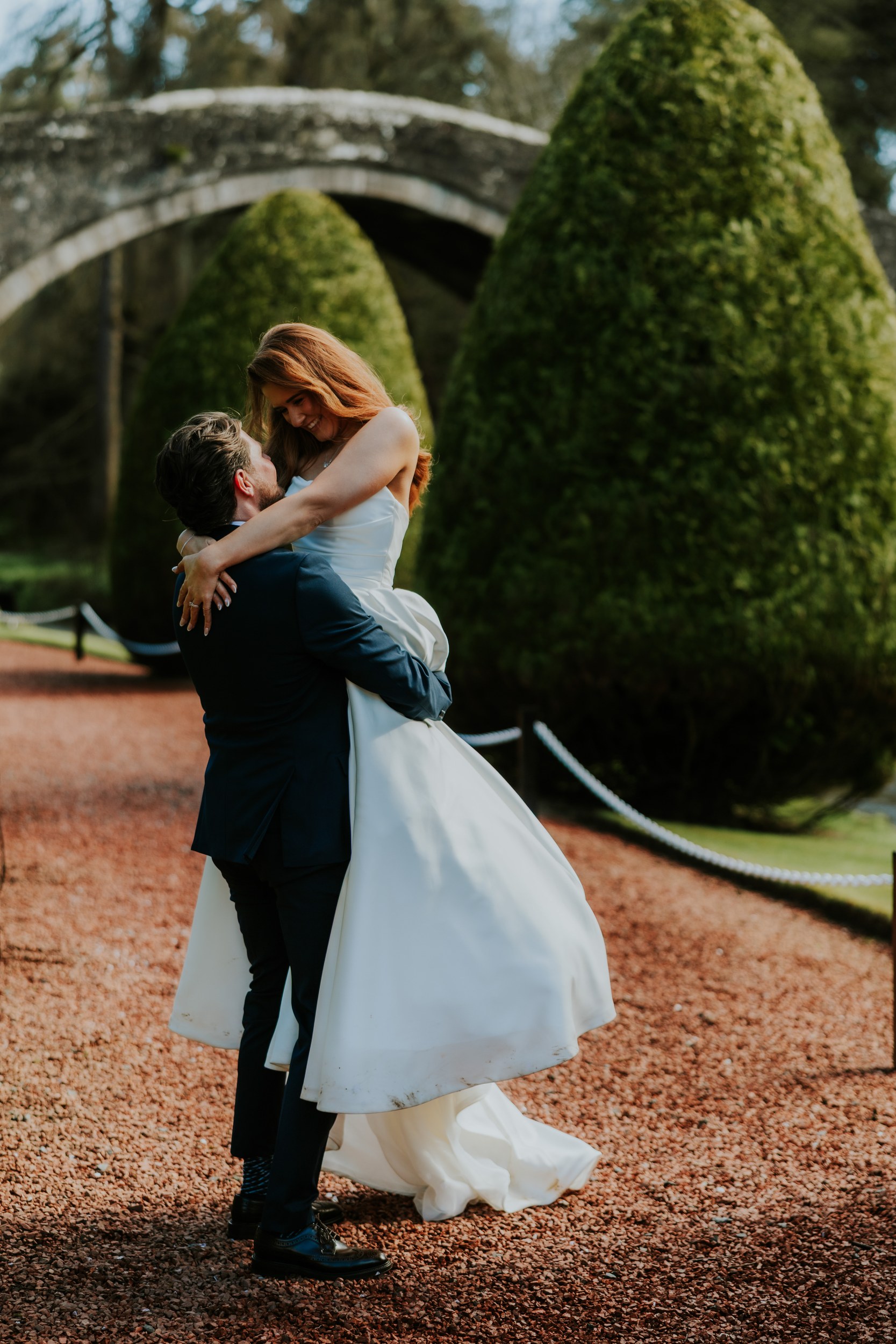 Groom lifts bride in white dress beneath a stone bridge and cone-shaped yew trees.
