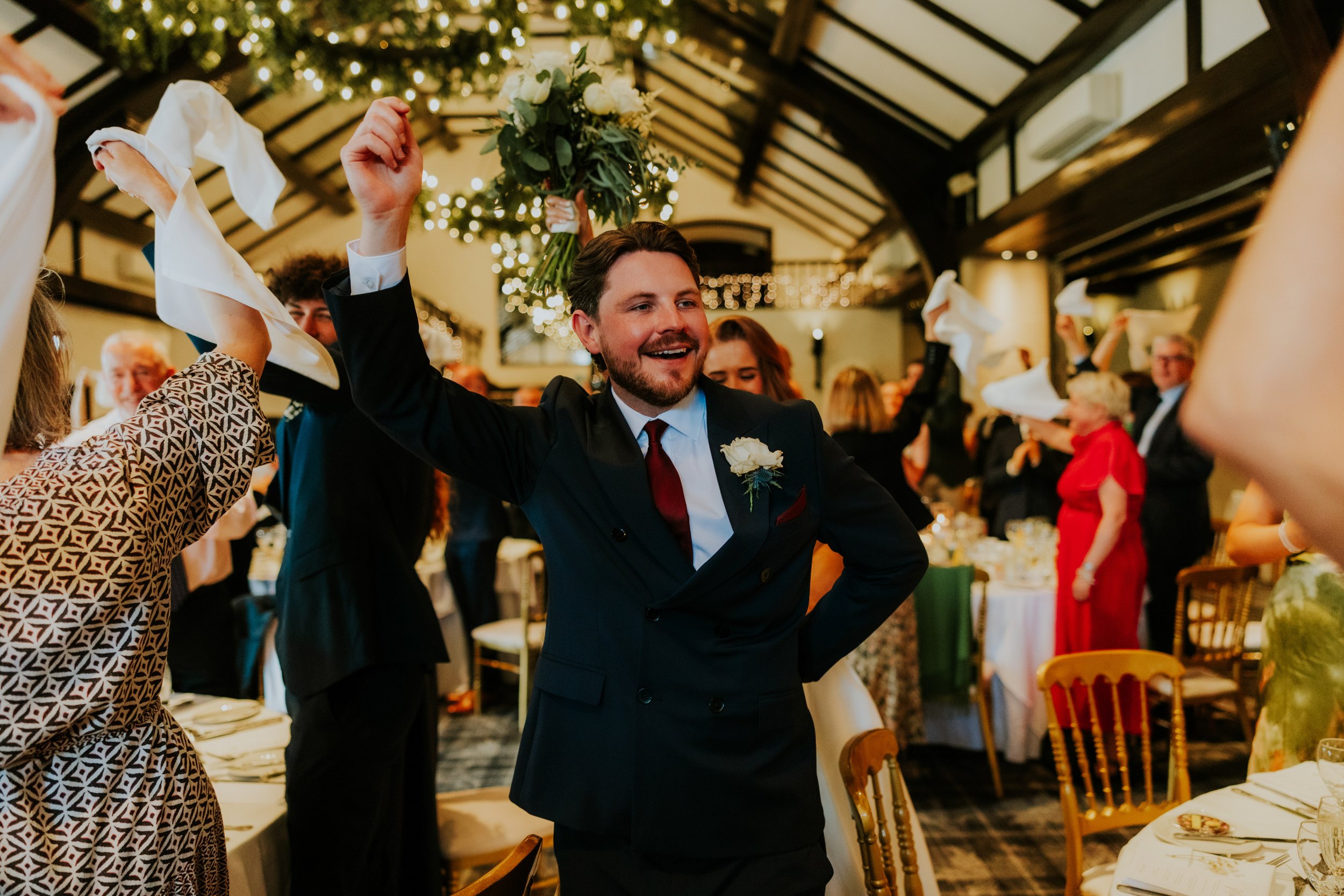 Groom smiling with arm raised as guests wave napkins in a timber‑beamed reception room with fairy lights.