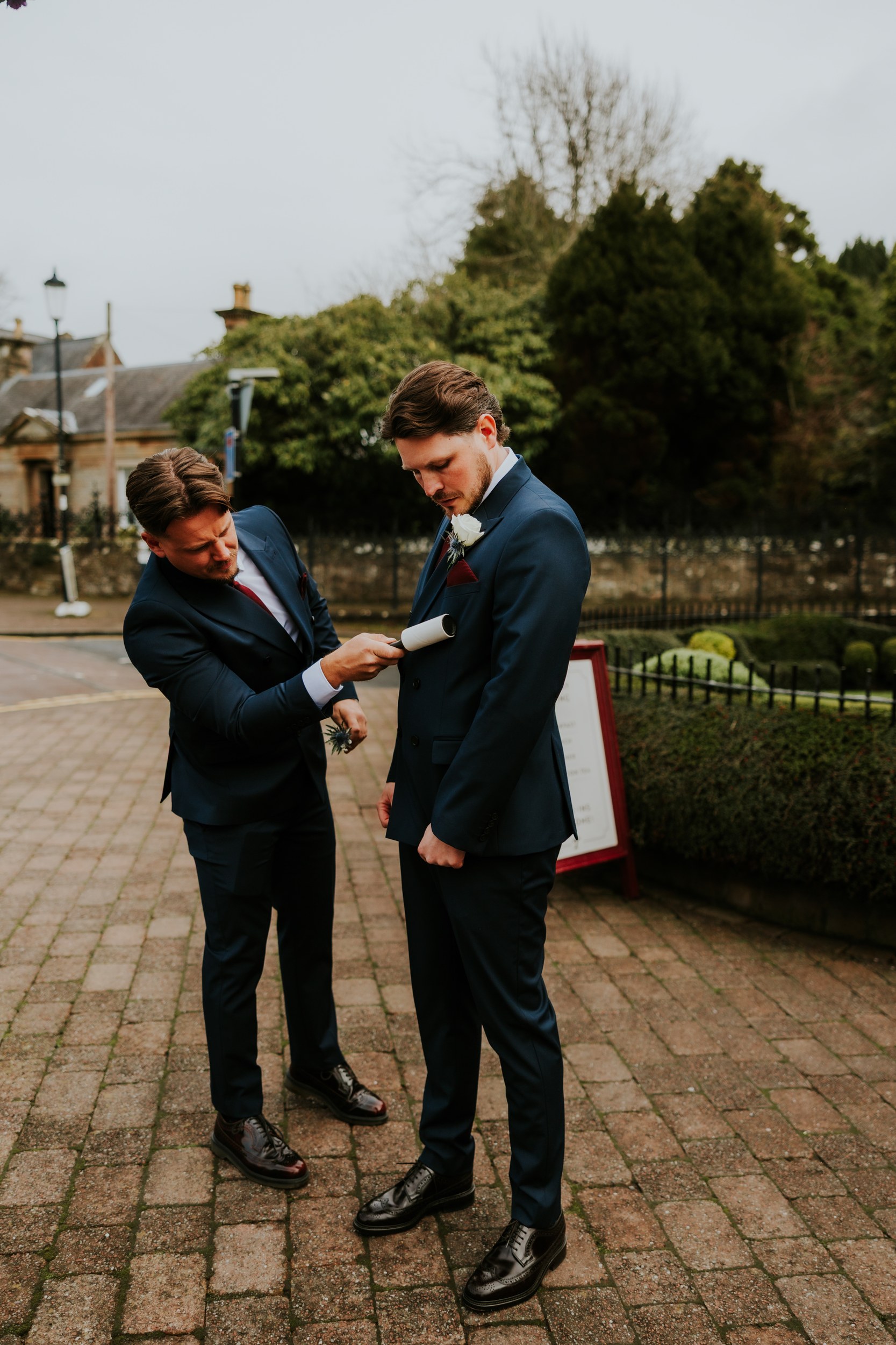 Groomsman lint-rolling the groom's navy suit jacket outside the Brig o' Doon House Hotel.