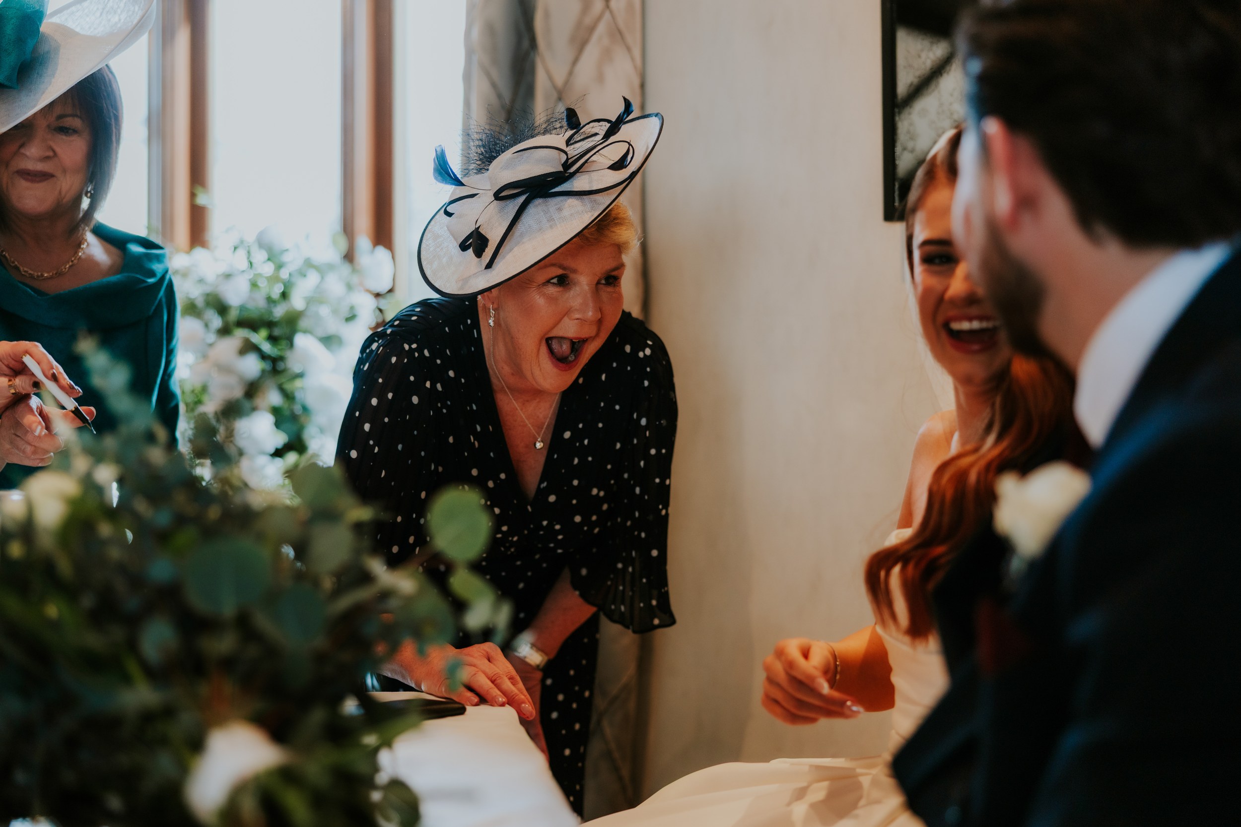 Guest in white-and-navy fascinator reacts as bride and groom smile at the signing table with flowers.