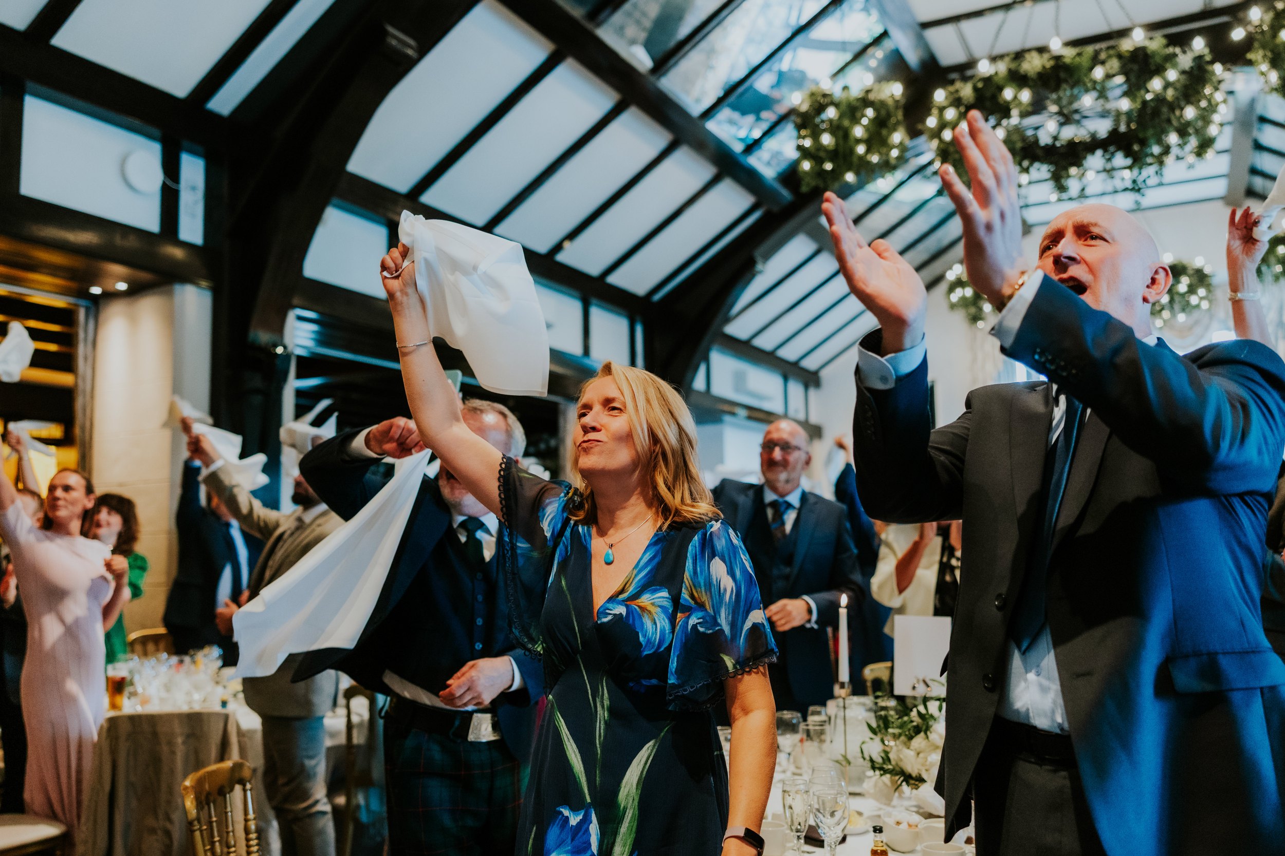 Guests in a timber‑beamed reception room waving white napkins under hanging greenery and lights.