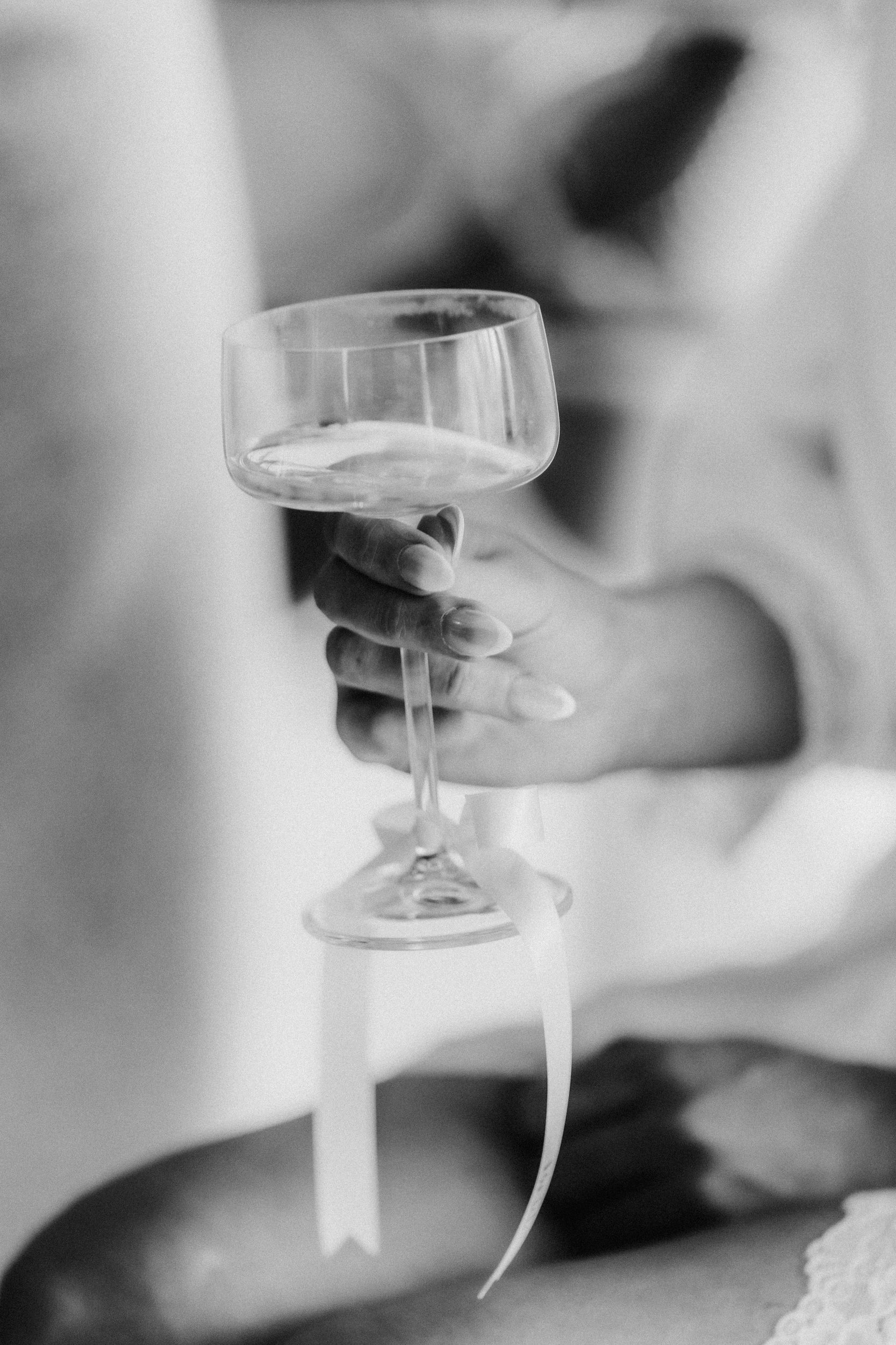 Hand holding a coupe glass with a ribbon on the stem, shallow focus and a blurred figure behind.