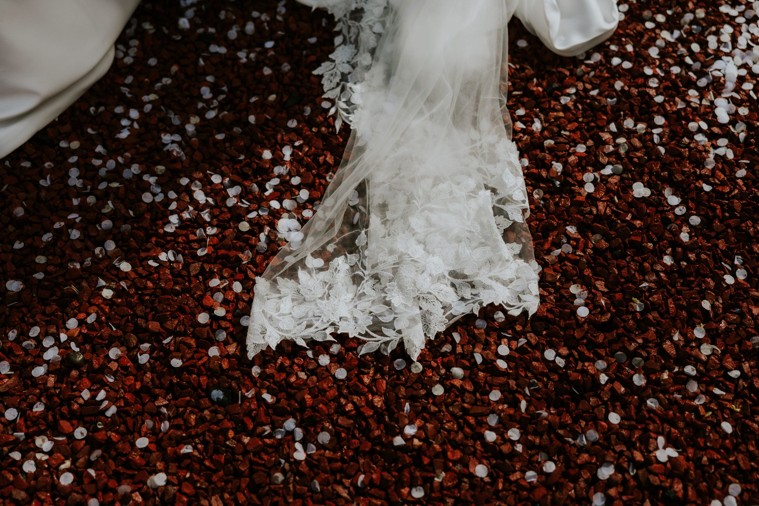 Lace wedding veil and gown hem on red gravel scattered with white confetti.