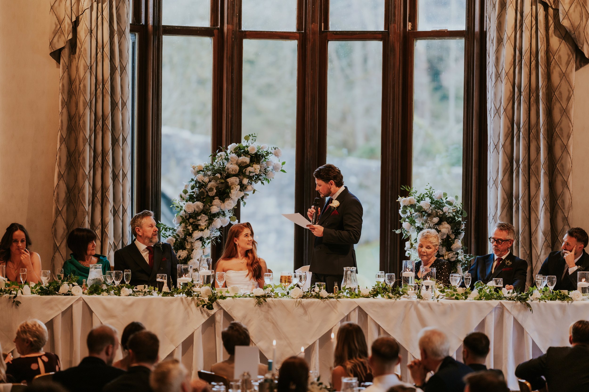 Man delivering a wedding speech as bride and family sit at a decorated head table under tall windows.