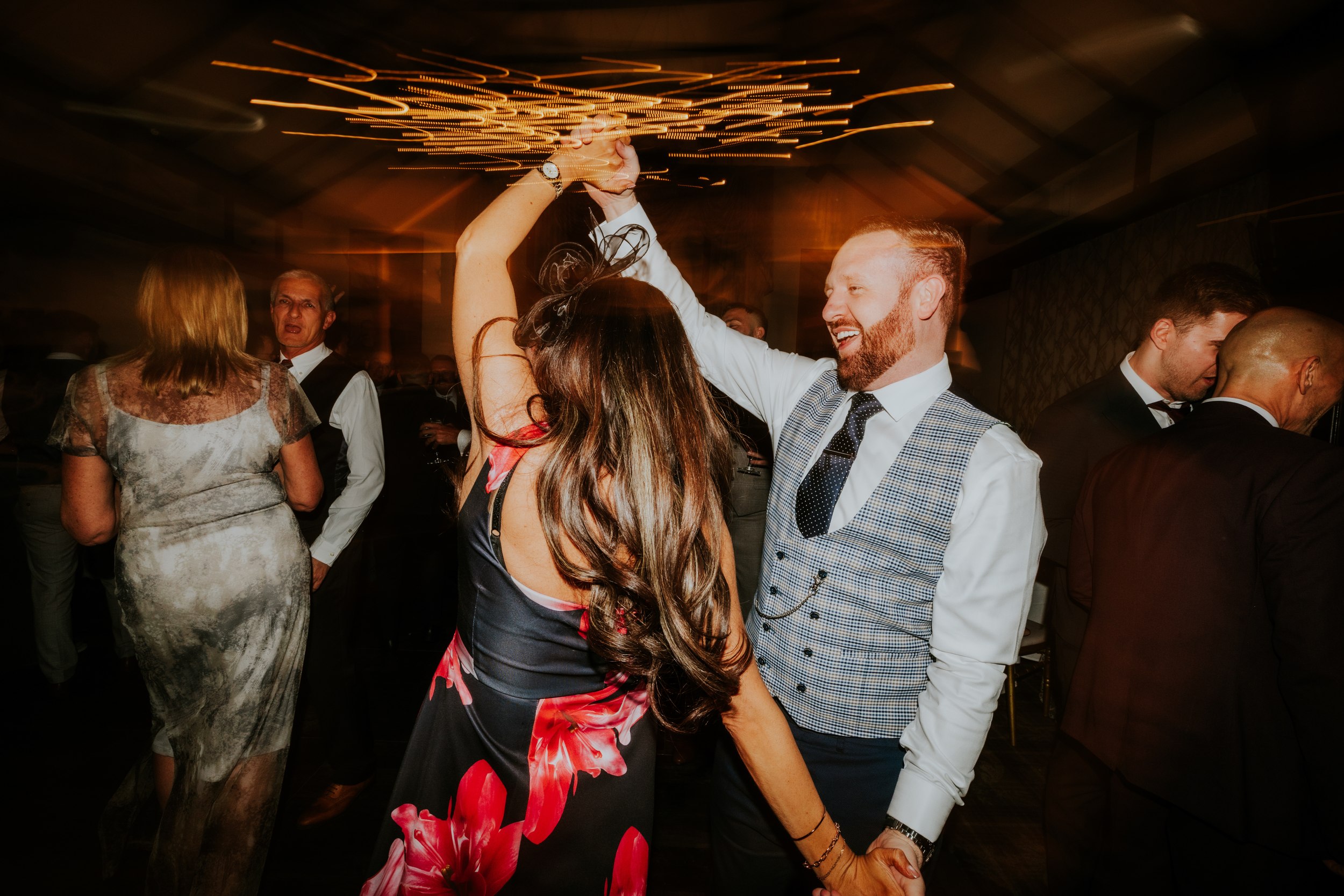 Man in checked waistcoat twirling a woman in a floral dress on the dancefloor with streaked overhead lights.