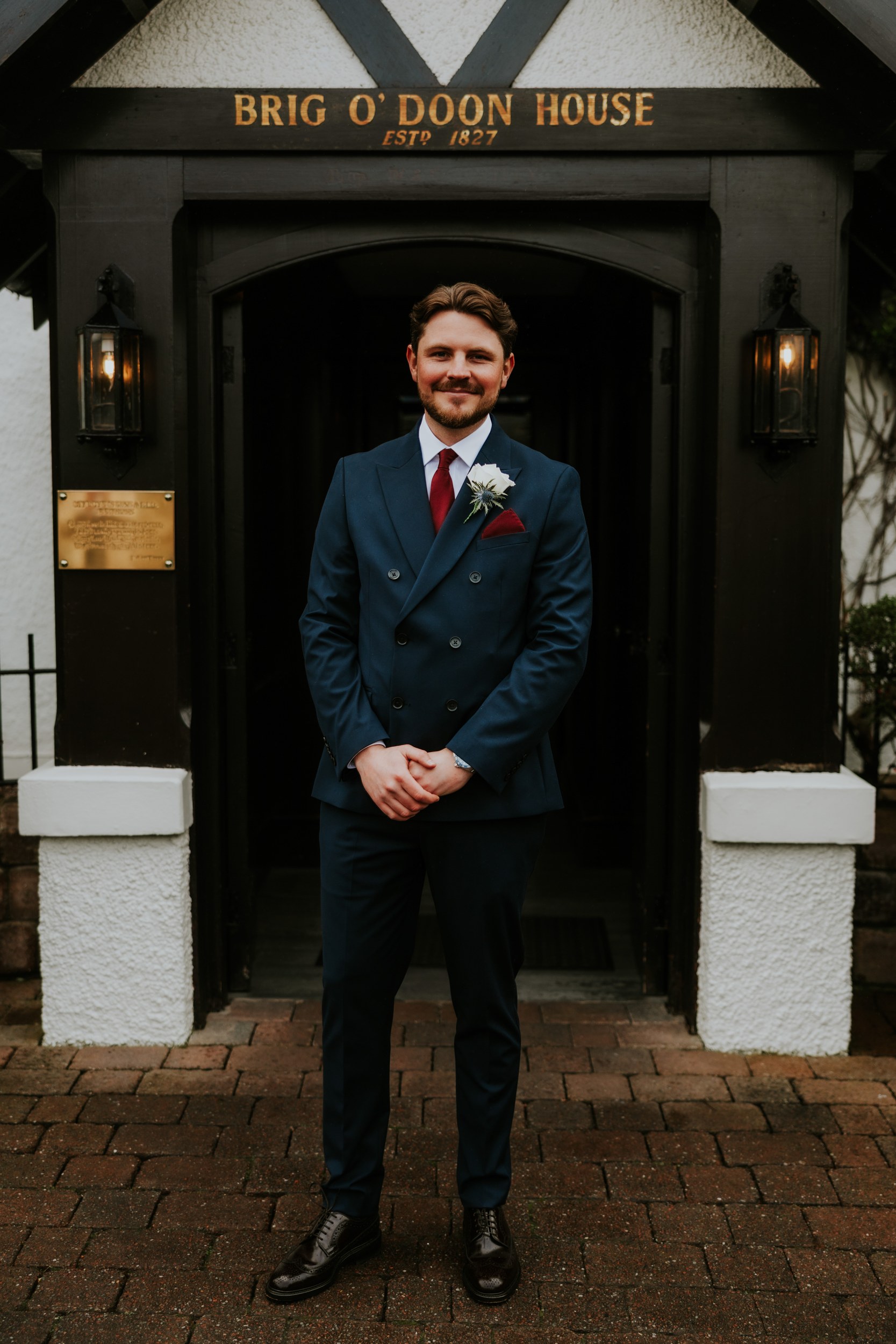 Man in navy suit with white boutonniere standing beneath the 'Brig O' Doon House' entrance sign.
