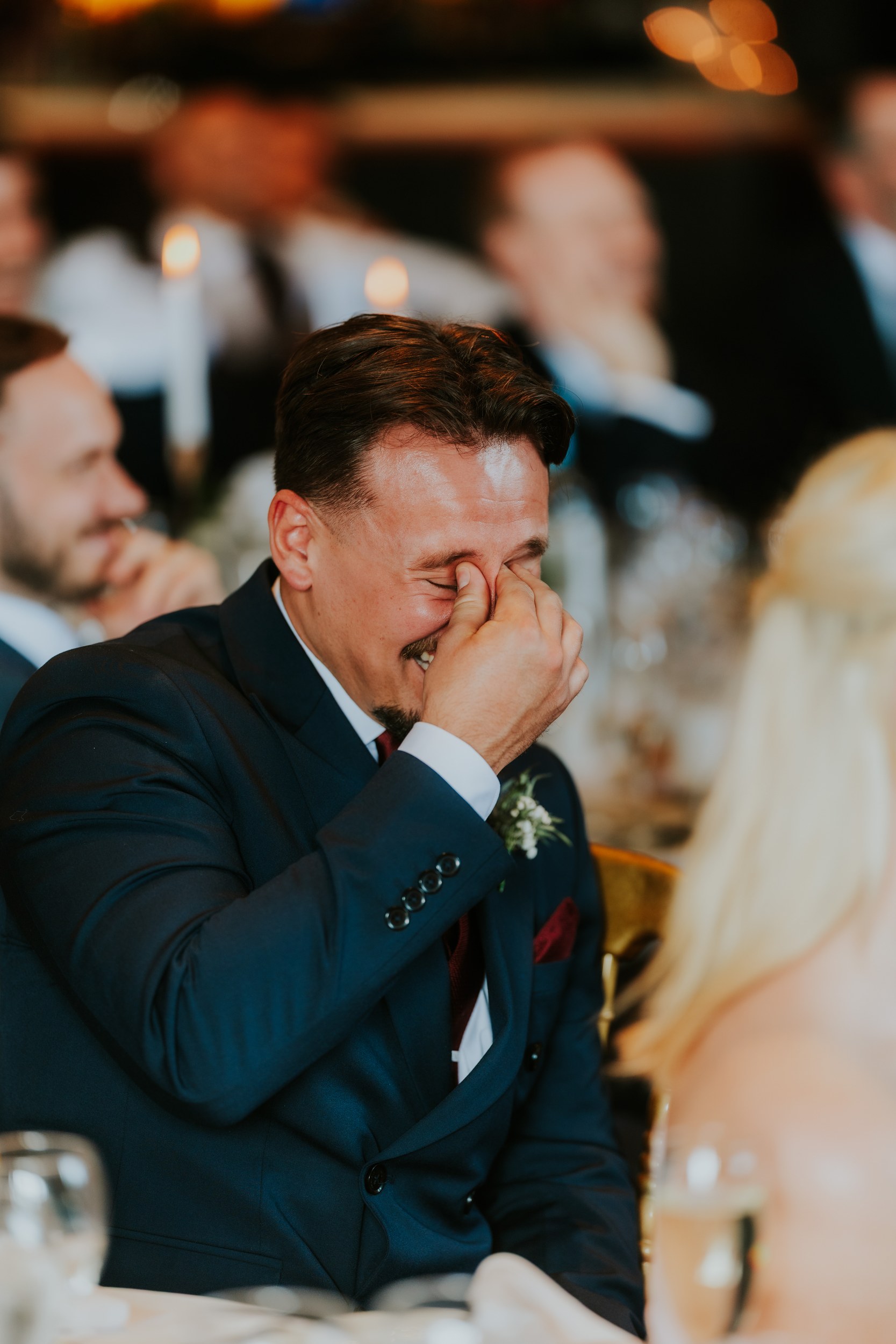 Man in suit laughing with hand over his face at a wedding reception, blurred guests and candles behind him.