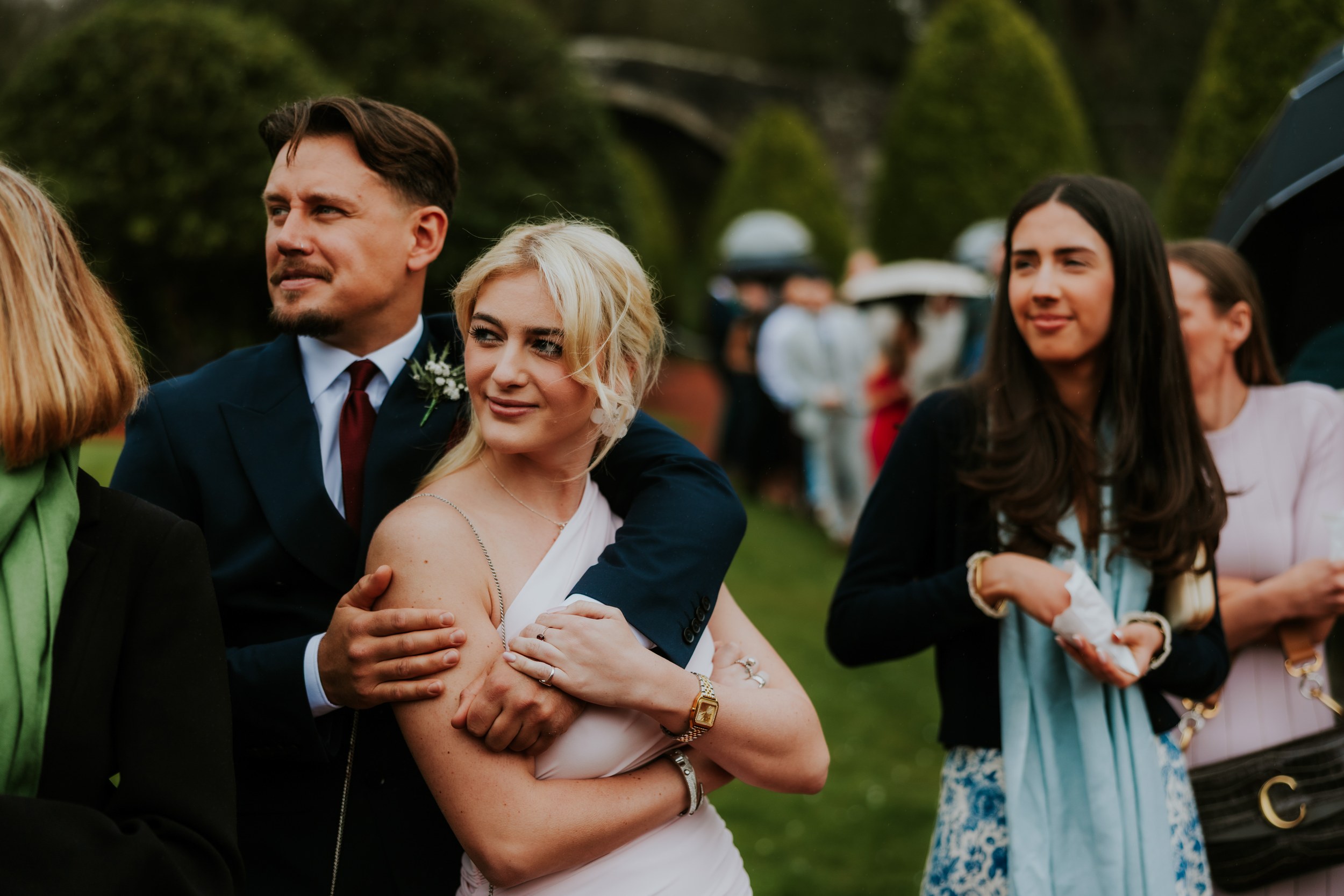 Man in suit with boutonniere embraces a blonde woman in a pale dress outdoors, guests and stone bridge arch blurred behind them.