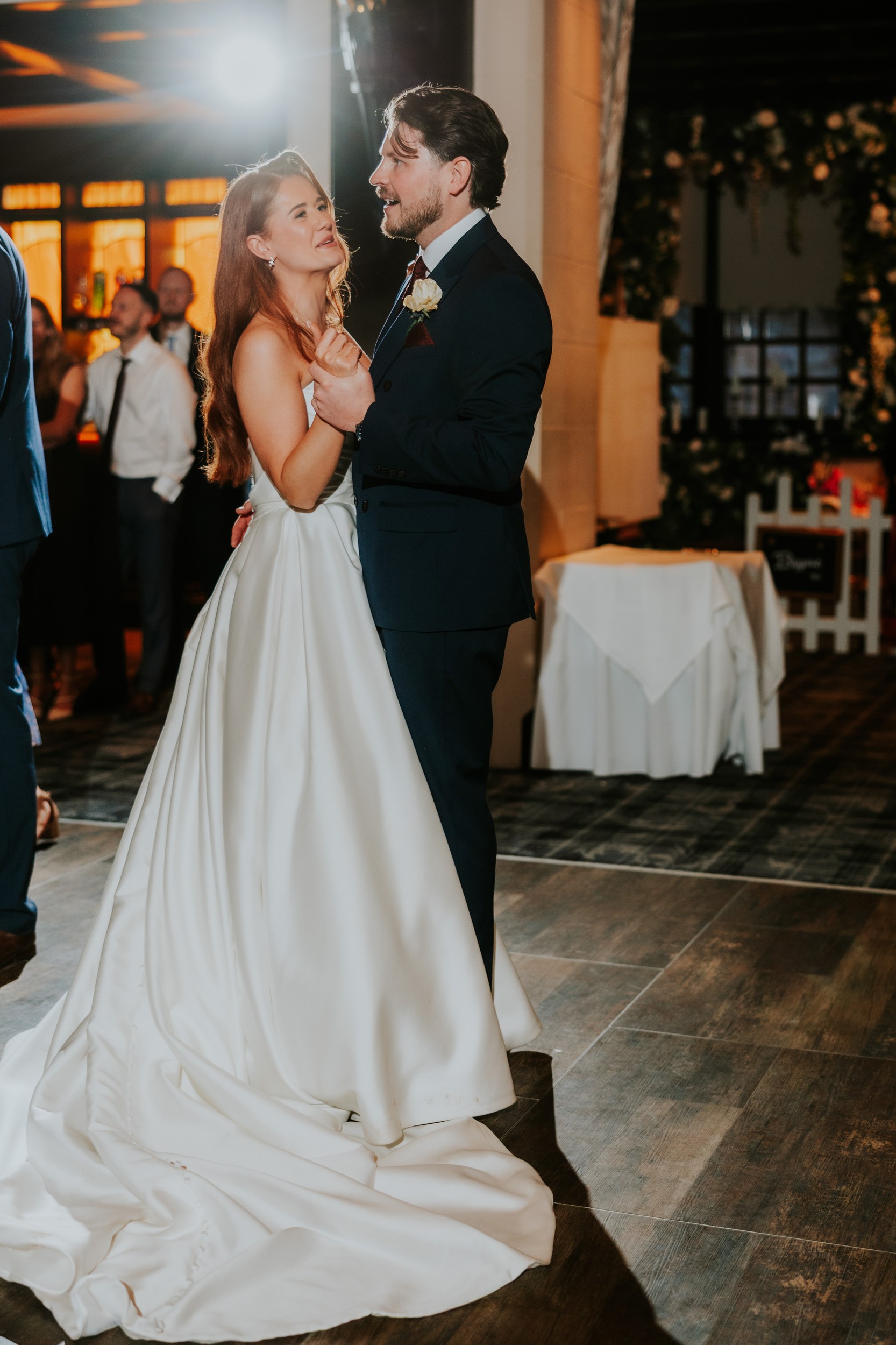 Newlyweds dancing at reception, bride in white satin gown and groom in dark suit with boutonniere.