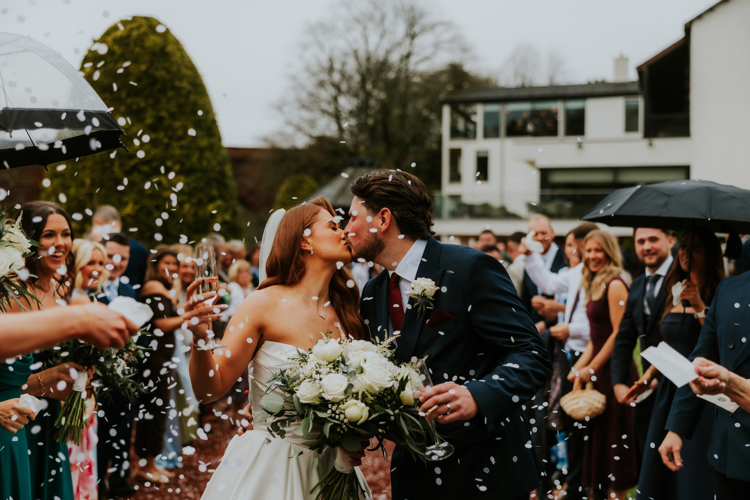 Newlyweds kiss as white confetti falls outside Brig o’ Doon House Hotel, bride holding bouquet with guests and umbrellas behind them.