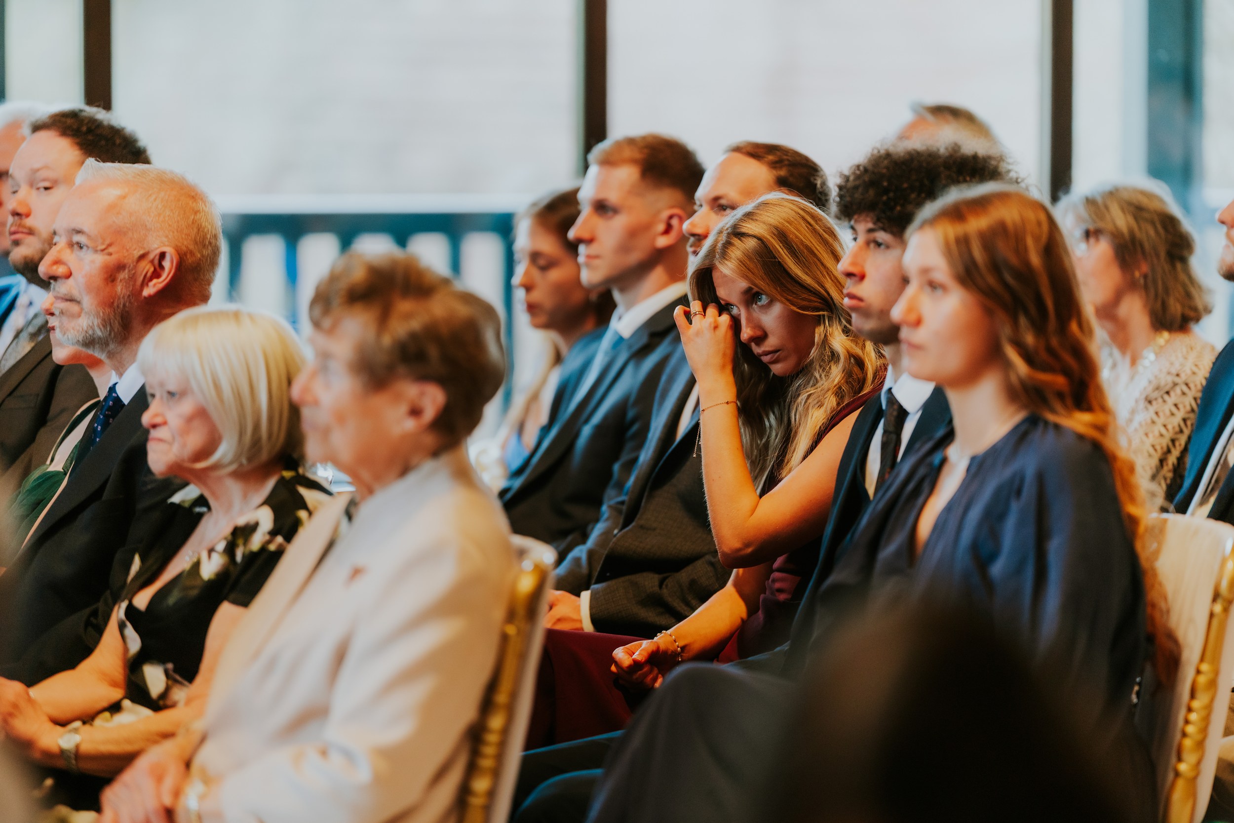 Seated wedding guests watching a ceremony, a woman wipes a tear while others sit in suits and dresses.