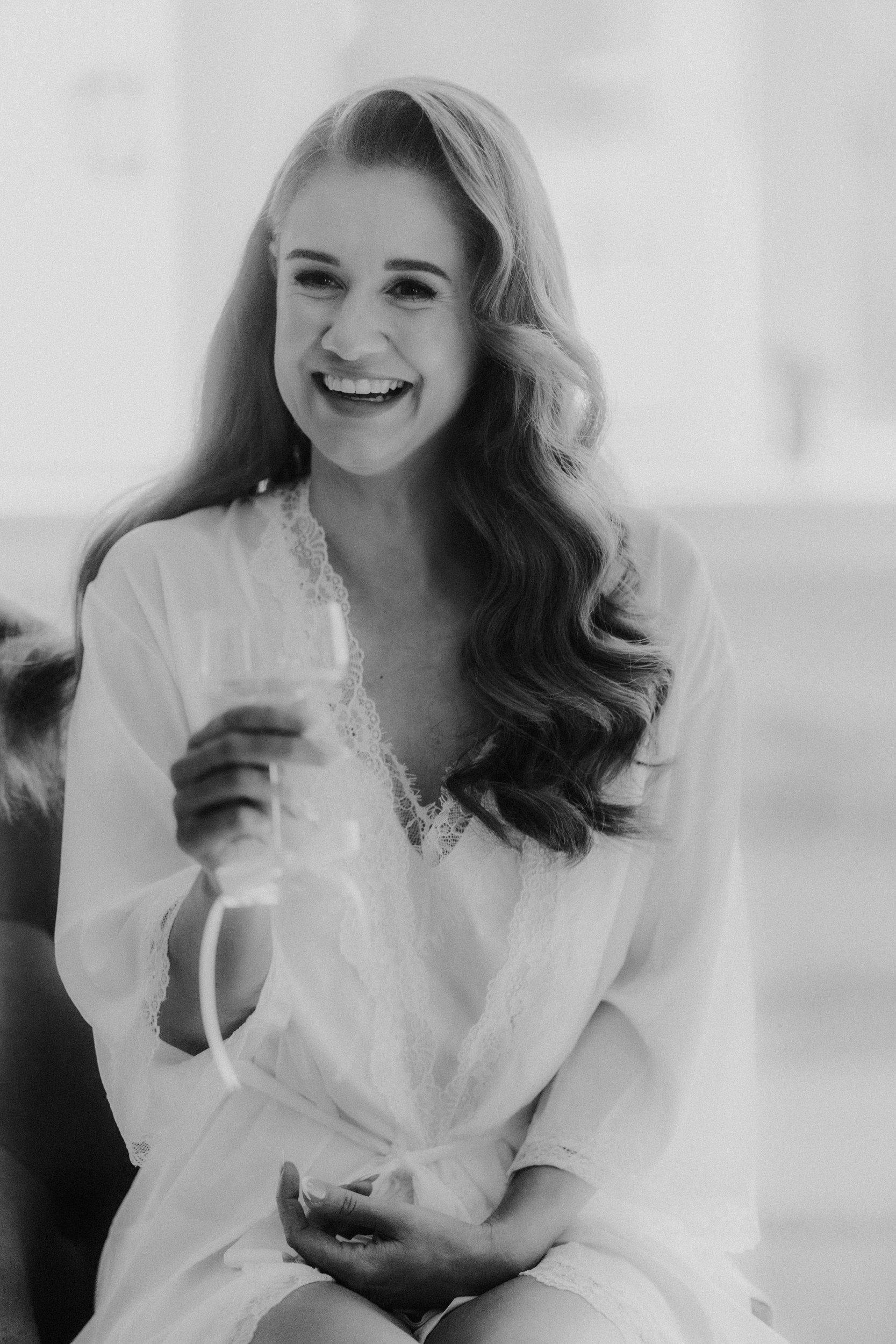 Smiling bride in a lace-trimmed robe holding a champagne flute.