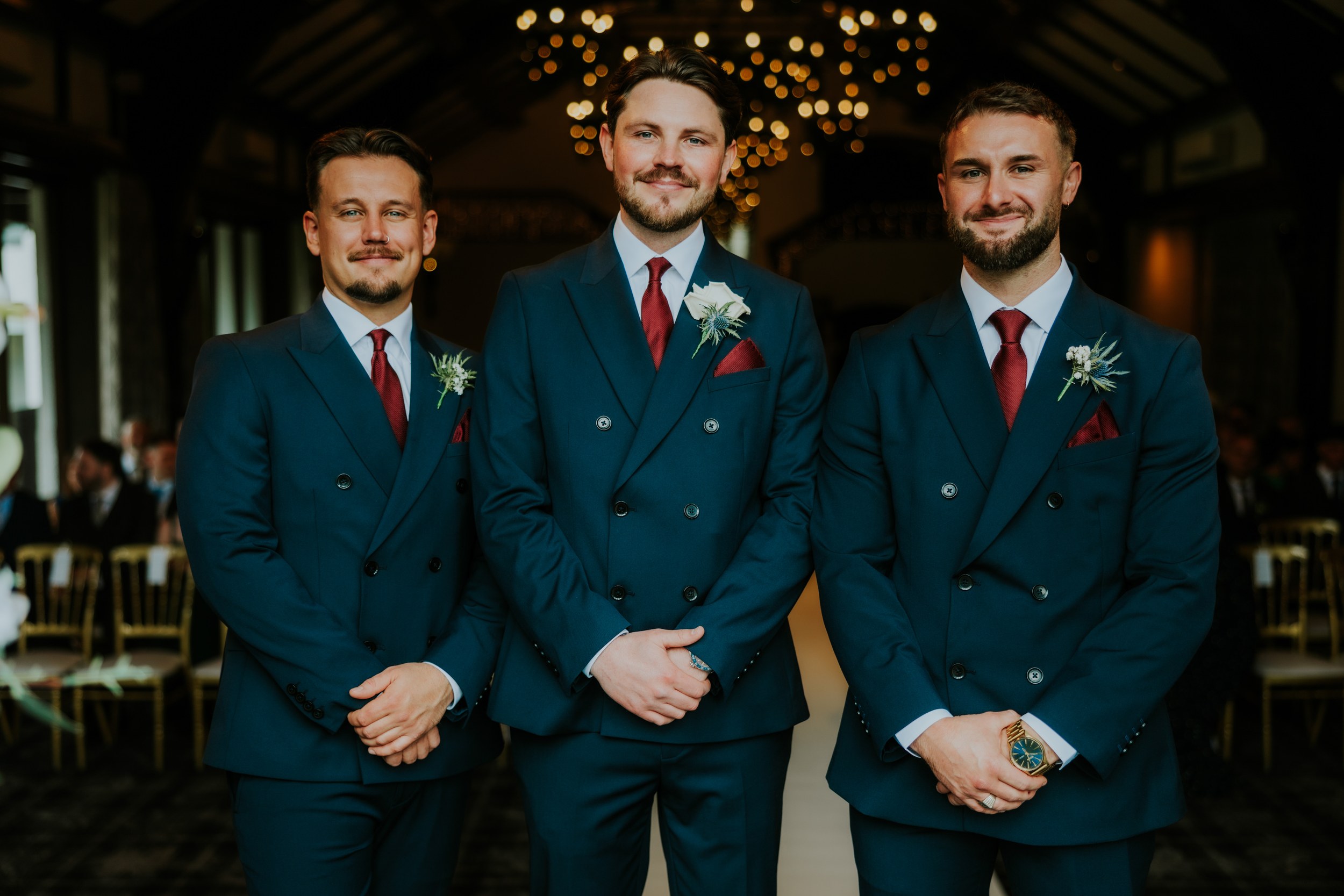 Three groomsmen in dark suits with burgundy ties and boutonnieres in a chandelier-lit ceremony room.