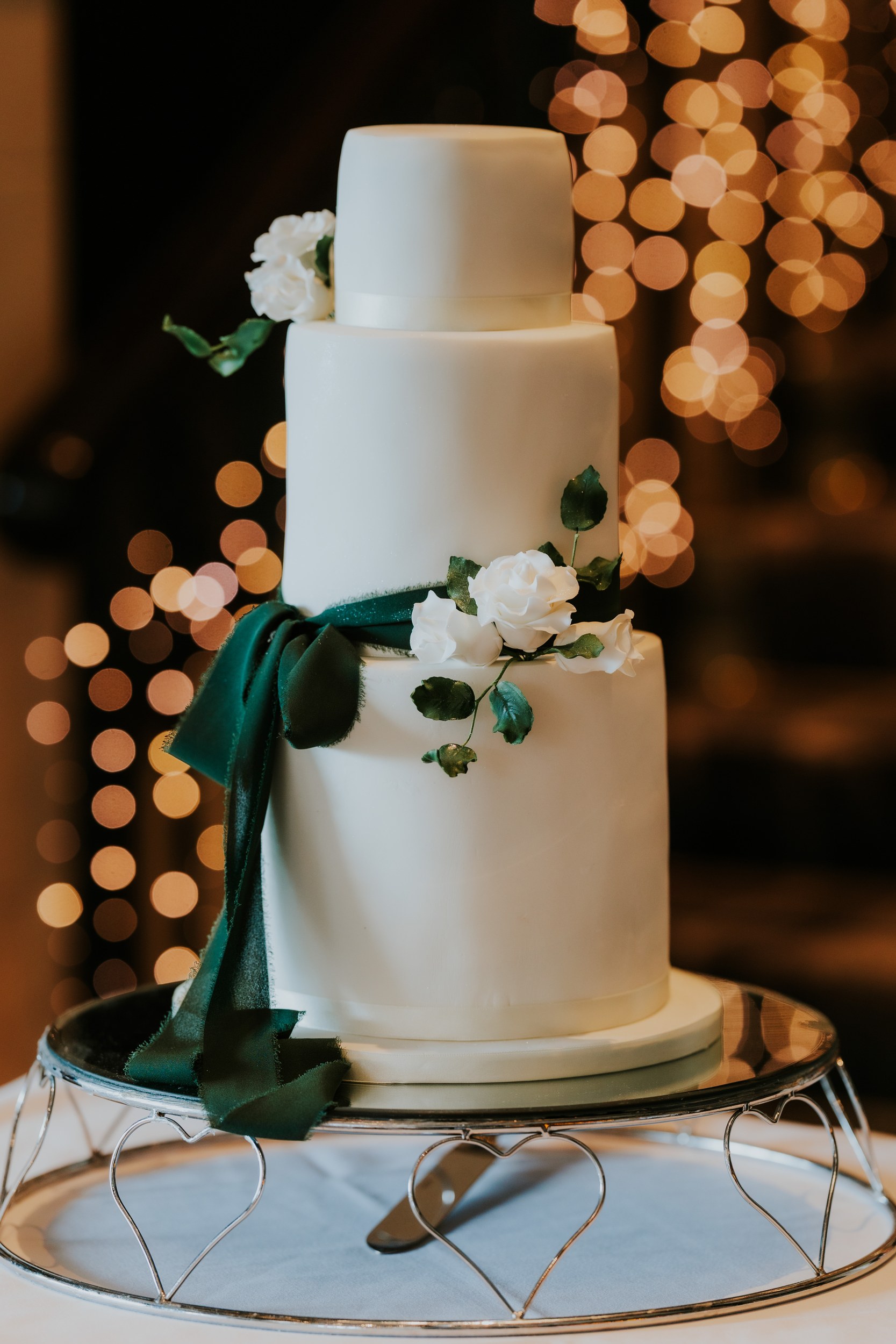 Three-tier white wedding cake with green ribbon and white roses on a silver stand, warm fairy lights blurred behind.