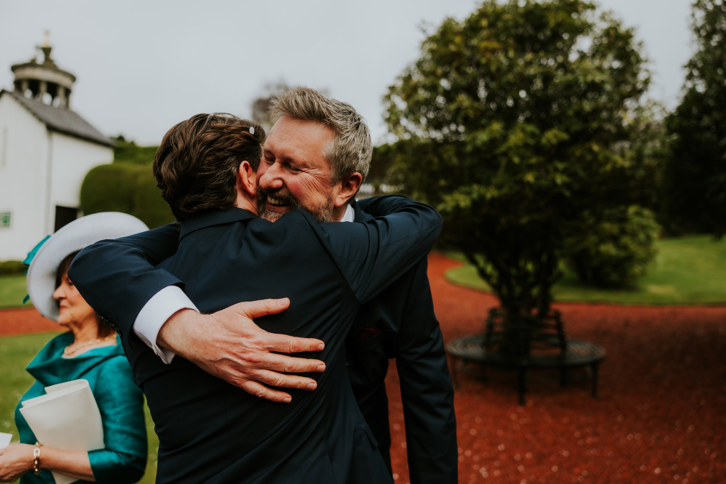 Two male wedding guests embrace outdoors; older grey‑haired man smiles, woman in teal and white hat nearby.