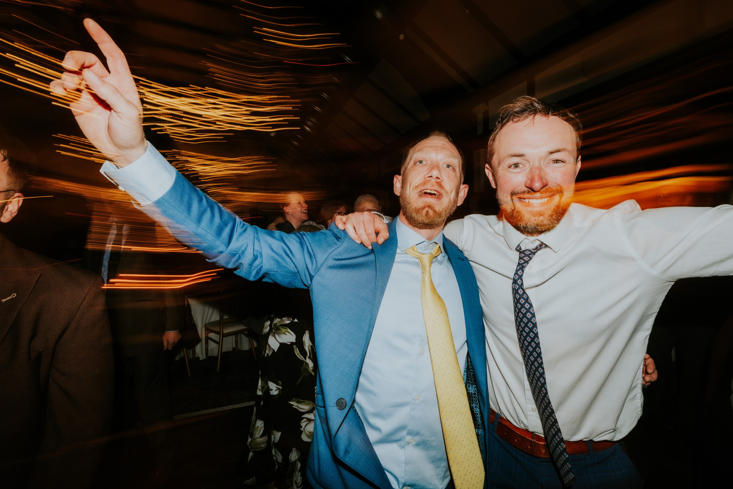 Two men at a wedding reception smiling with arms round each other, warm light trails streaking behind them.