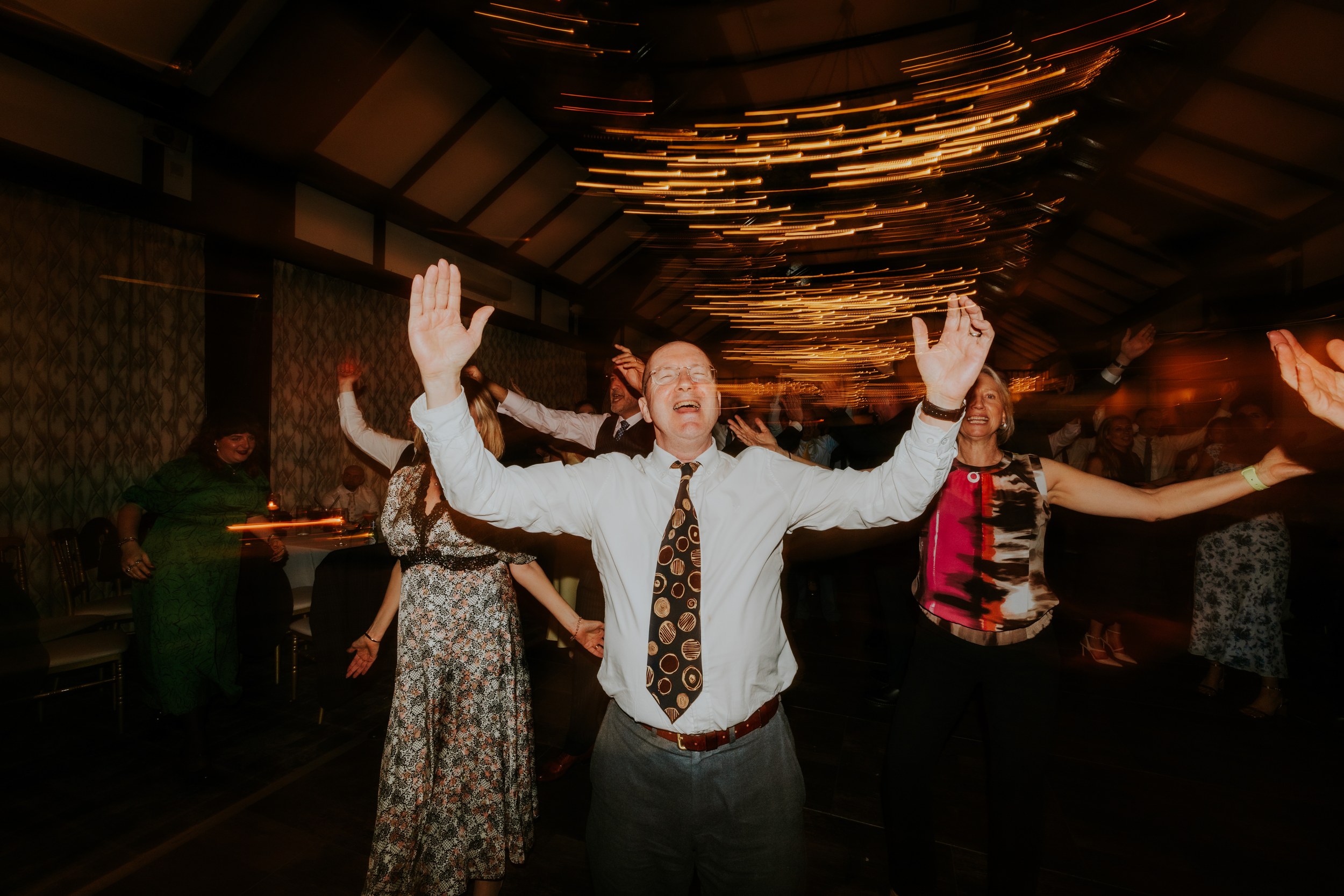 Wedding guests dancing with arms raised and streaked ceiling lights overhead.