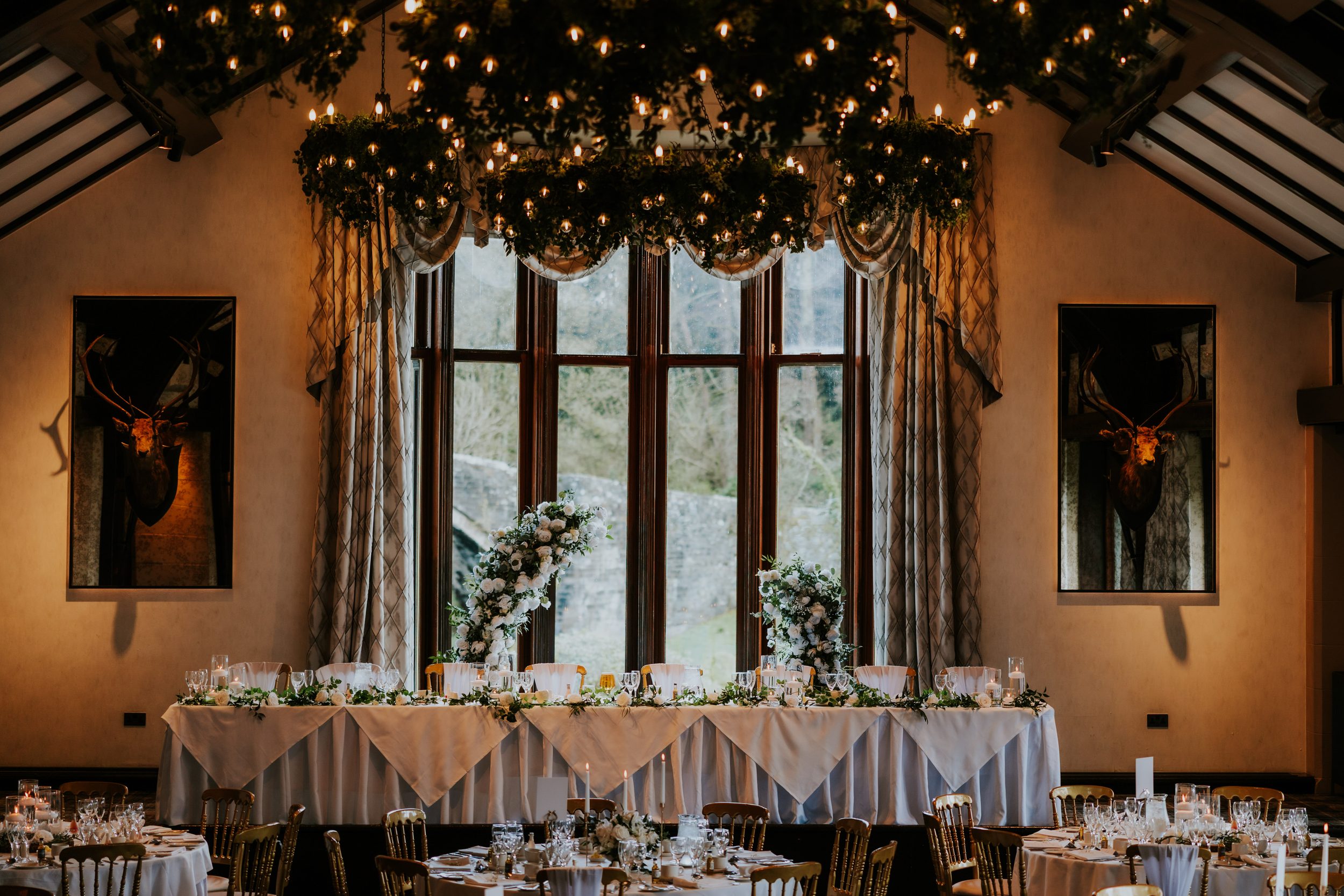 Wedding head table under a tall window with floral arrangements, hanging foliage chandeliers and gold‑backed chairs.
