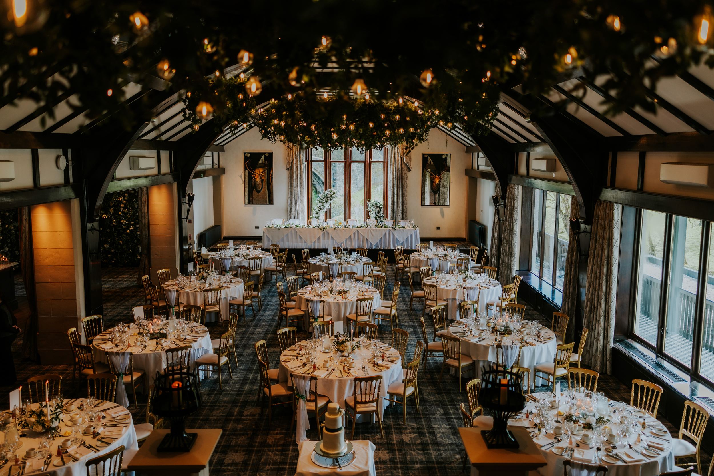 Wedding reception at Brig o’ Doon House Hotel with round tables, gold chairs, head table by tall windows and hanging greenery with lights.