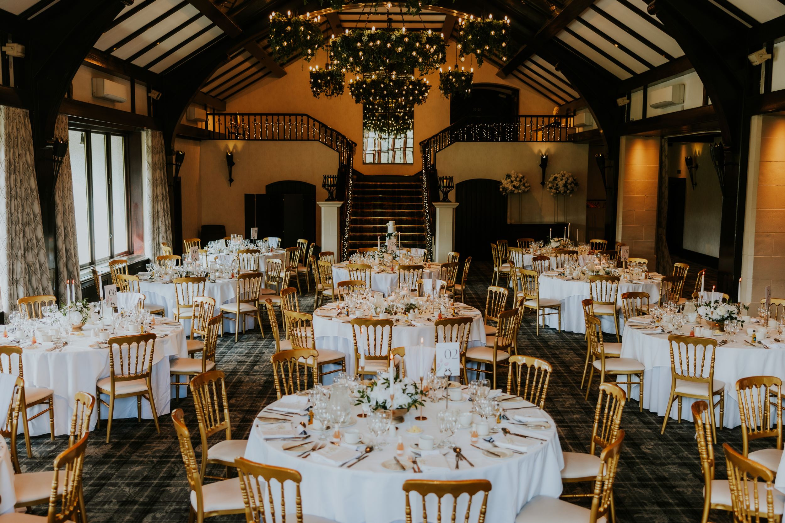 Wedding reception room with round tables, white linens, gold chairs, floral centrepieces and a lit staircase under chandeliers.