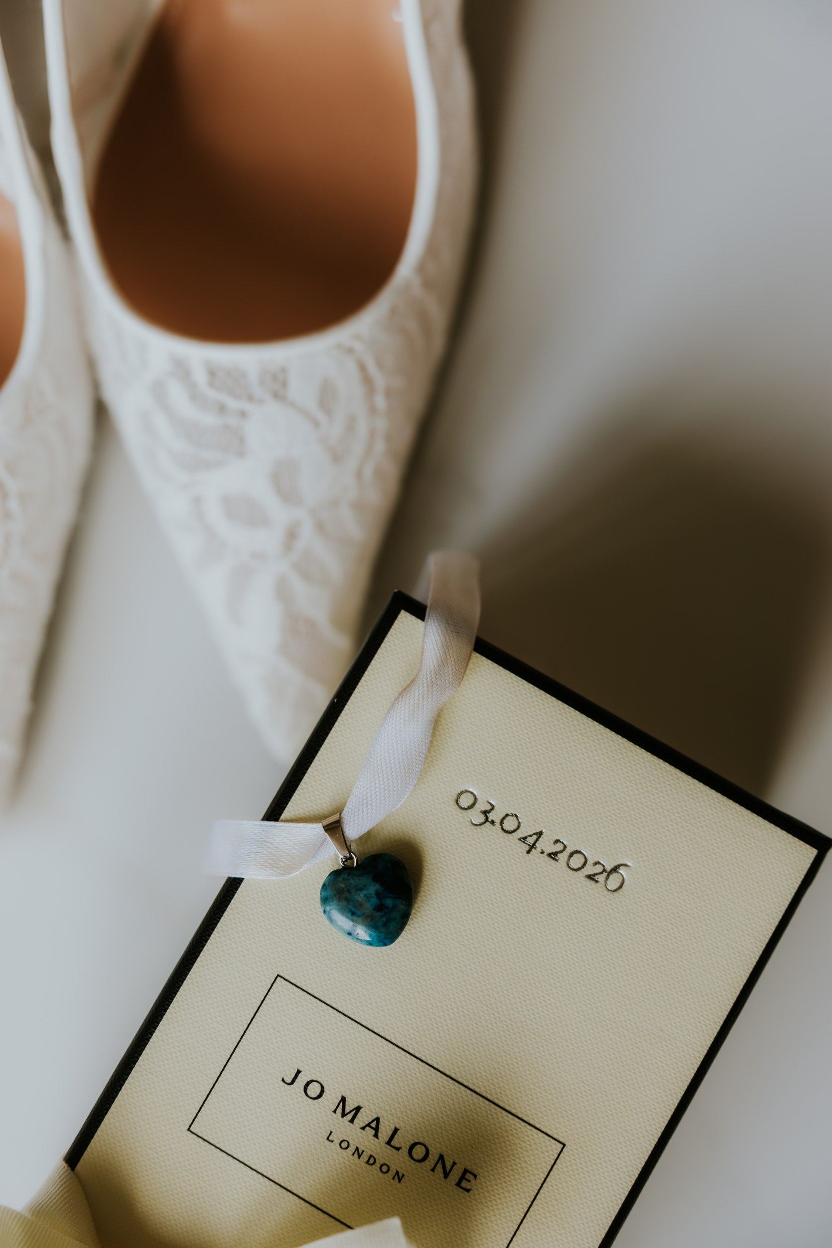 White lace wedding shoes with Jo Malone box dated 03.04.2026 and a ribboned turquoise heart pendant.