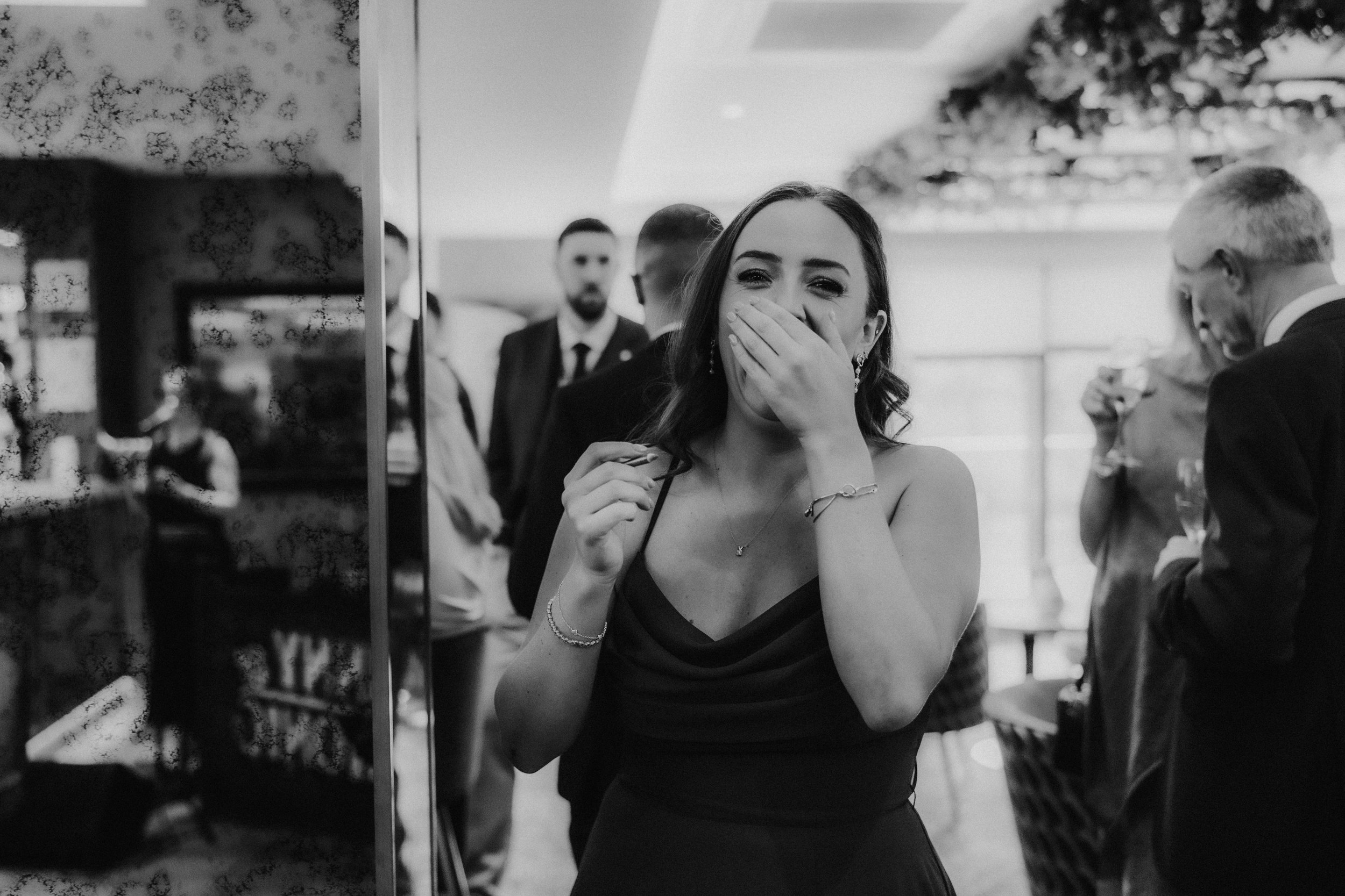 Woman in a dark dress covering her mouth laughing at a wedding reception, guests and drinks blurred behind her.