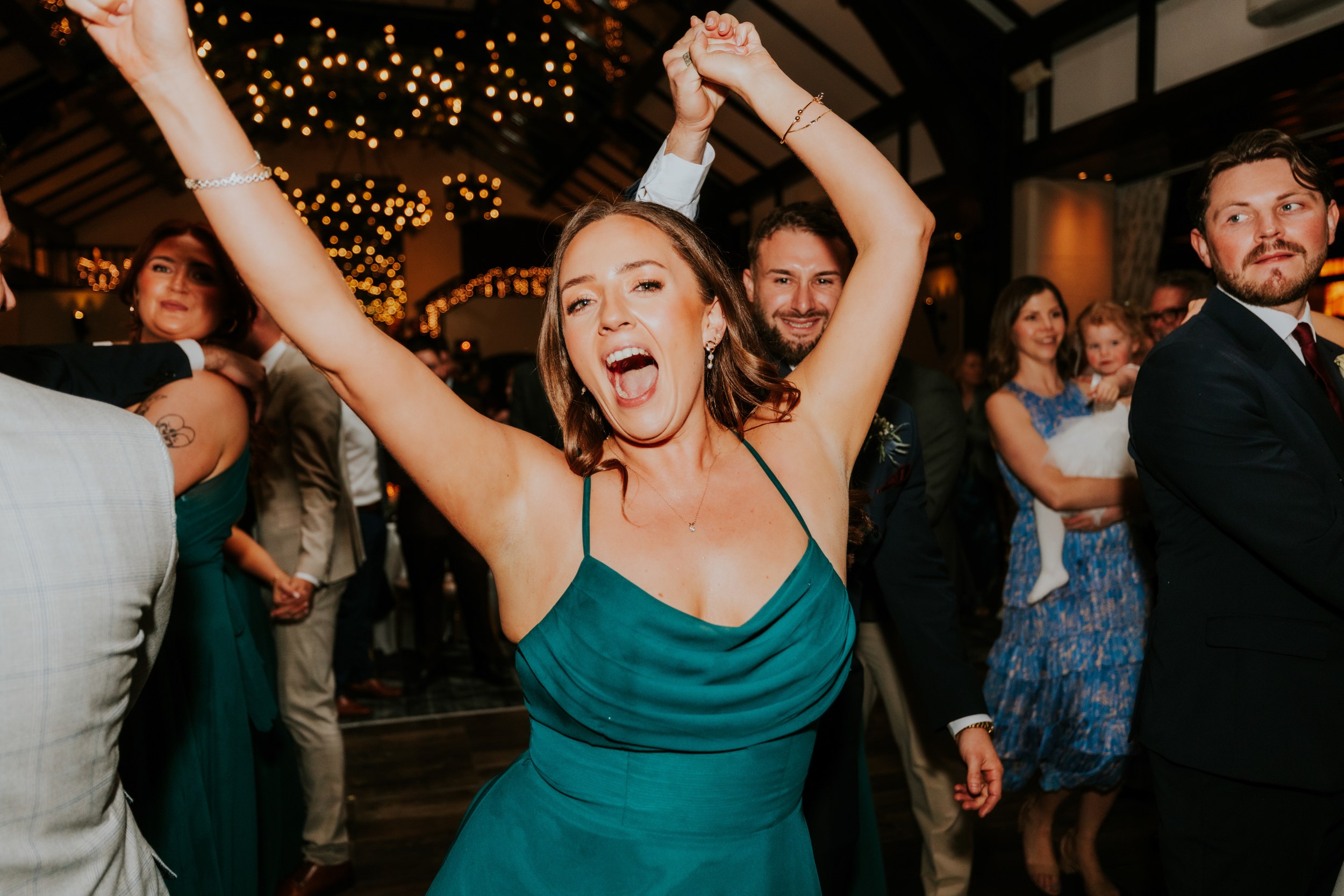 Woman in a teal dress cheering with arms raised on a wedding dance floor under fairy lights.