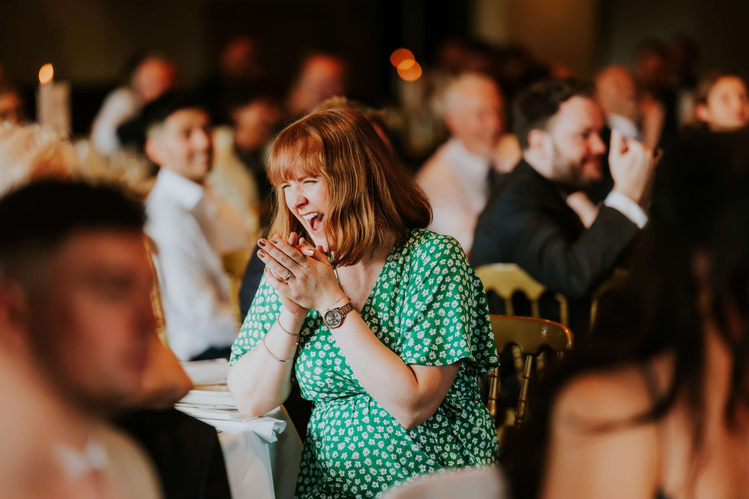 Woman in green floral dress laughing and clapping at a wedding reception, blurred guests behind her.