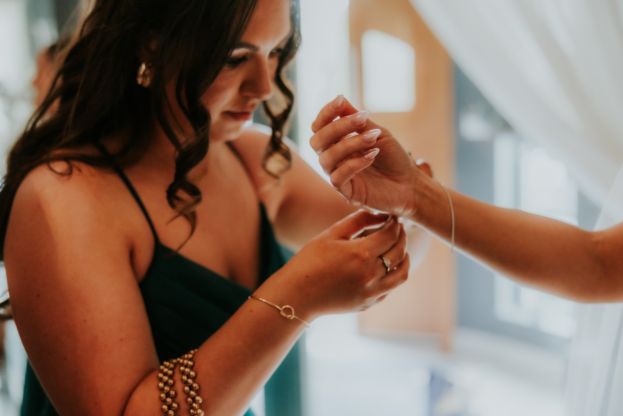 Woman with curled hair fastening a bracelet on another person's wrist during wedding preparations.
