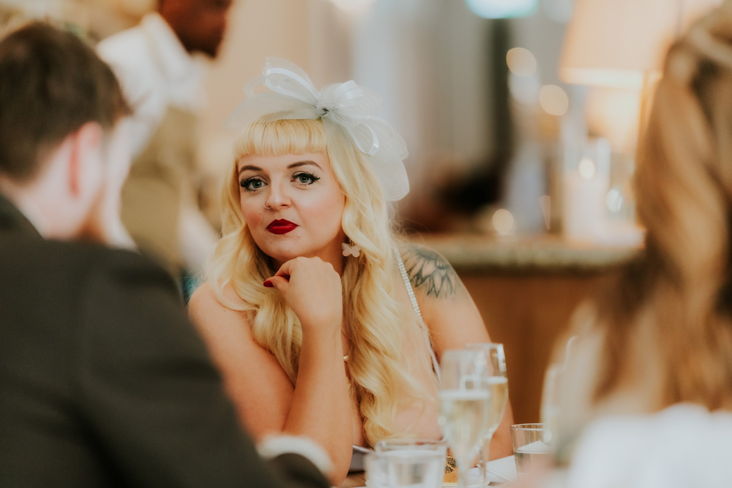 Citation Glasgow wedding photographer 70 Blonde woman with a white fascinator and red lipstick seated at a wedding reception, looking towards the camera.