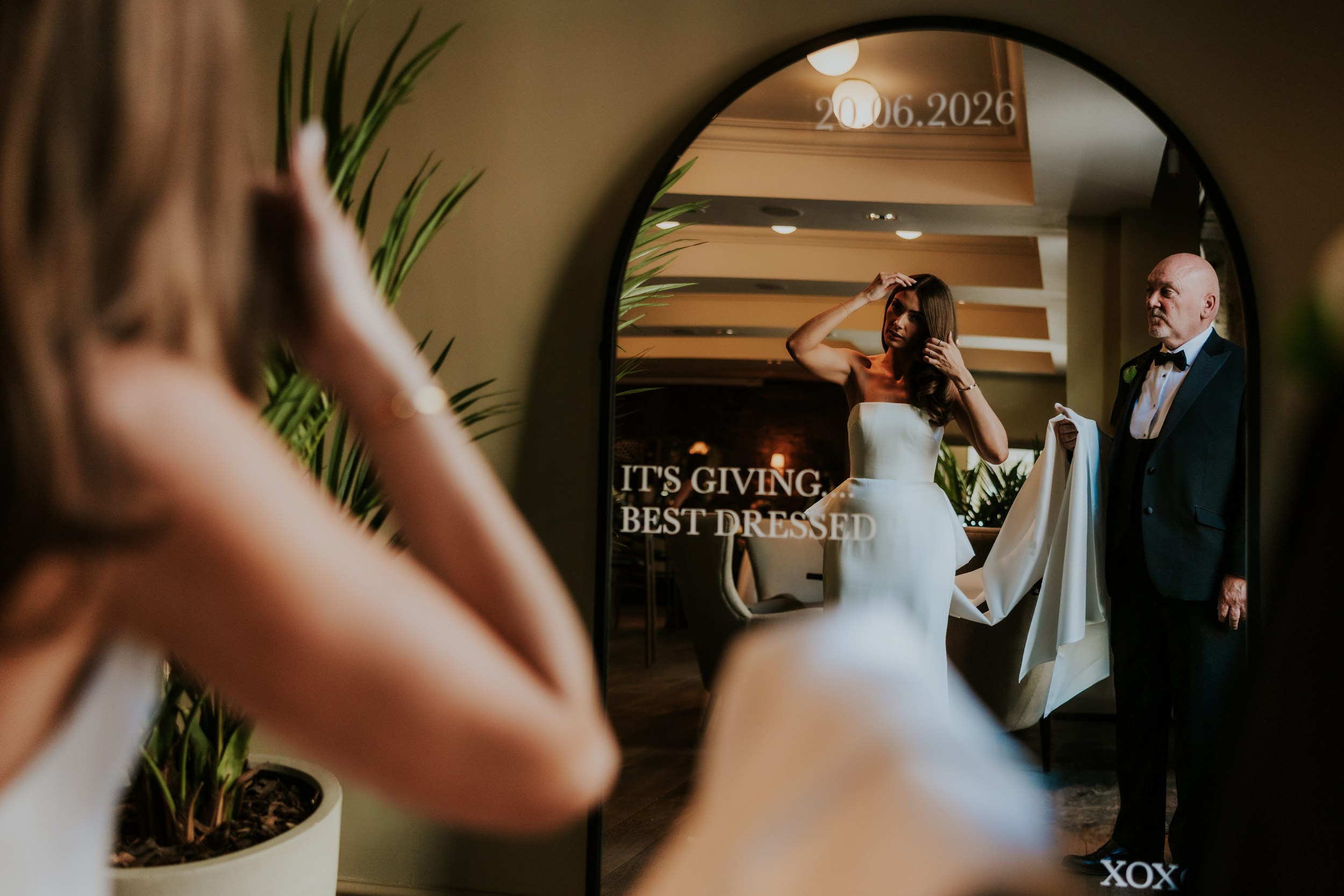 Citation Glasgow wedding photographer 23 Bride adjusting her hair in a mirror while a man in a tux holds her train inside the venue.