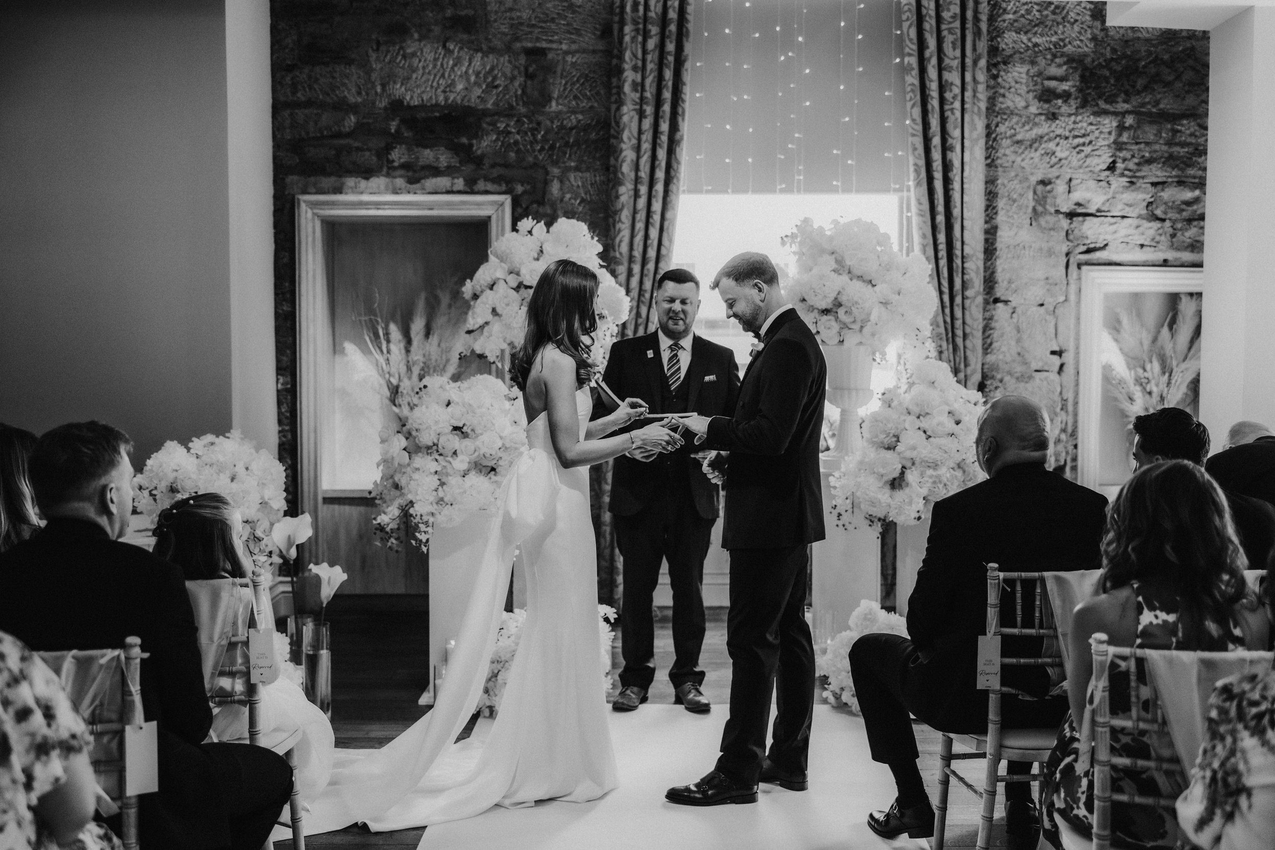 Citation Glasgow wedding photographer 33 Bride and groom exchange rings at an indoor ceremony with celebrant, large floral displays and seated guests.