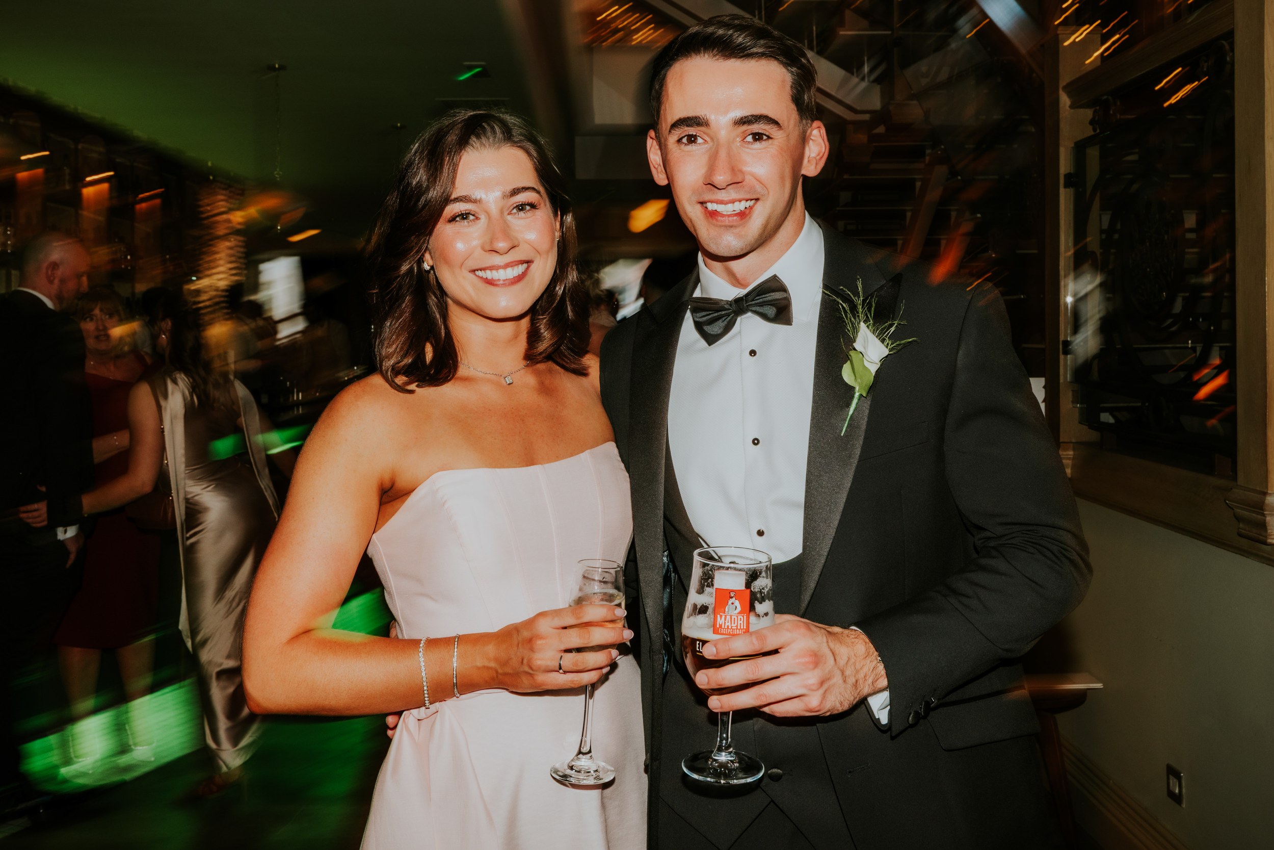 Citation Glasgow wedding photographer 88 Bride and groom in formalwear holding drinks at a busy reception, blurred guests and lights behind them.