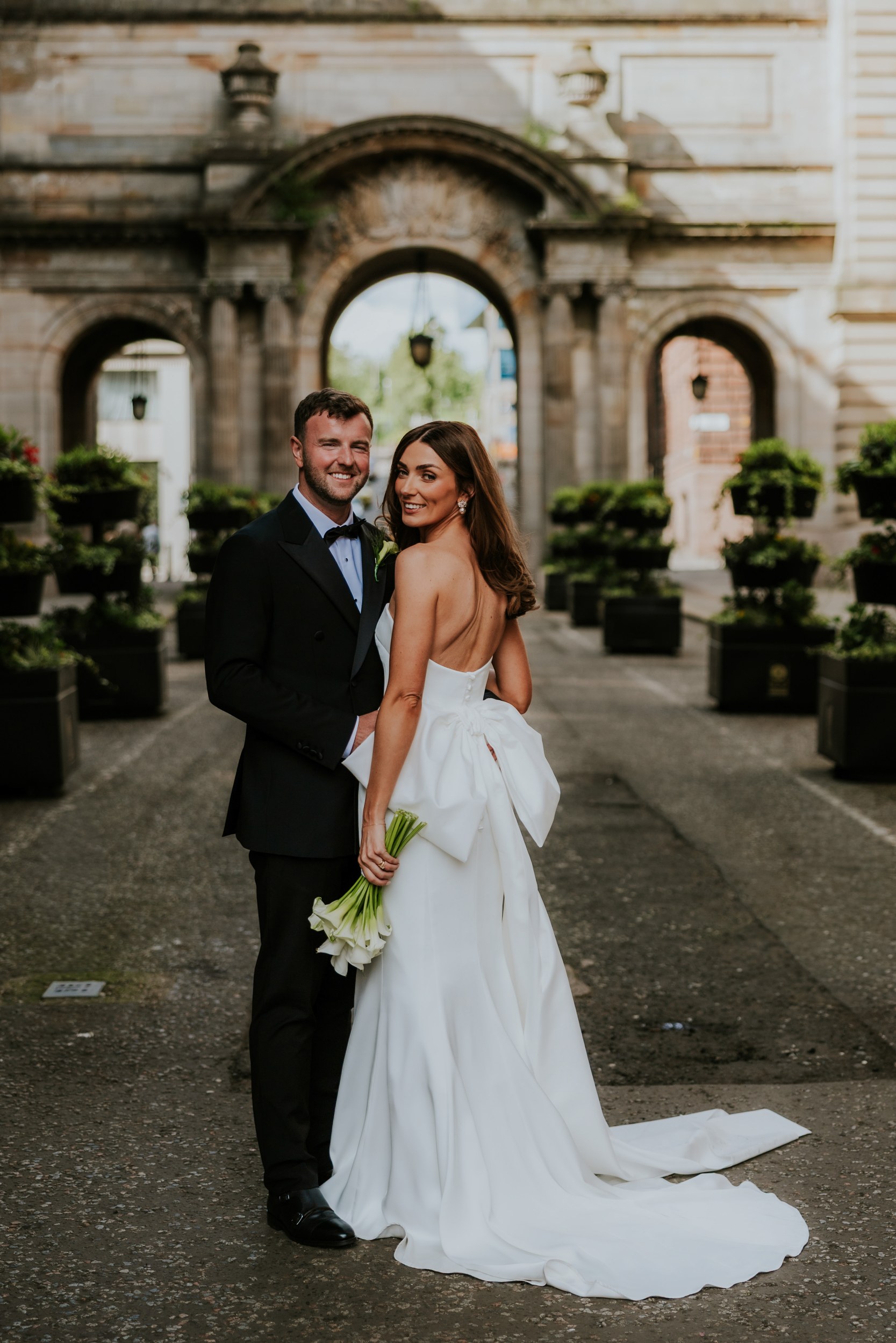 Citation Glasgow wedding photographer 50 Bride and groom in wedding attire stand under a stone archway outside Citation Glasgow, the bride holding calla lilies.