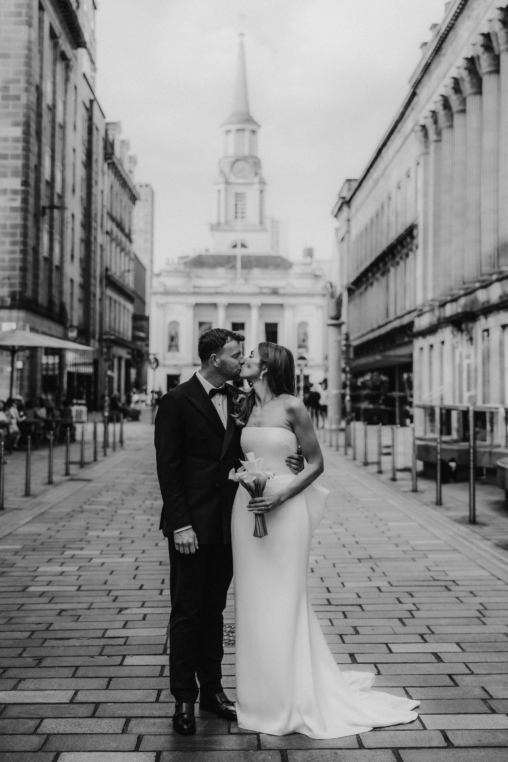 Citation Glasgow wedding photographer 58 Bride and groom kiss on a Glasgow street, bride in a strapless dress holding a bouquet, groom in a black suit.