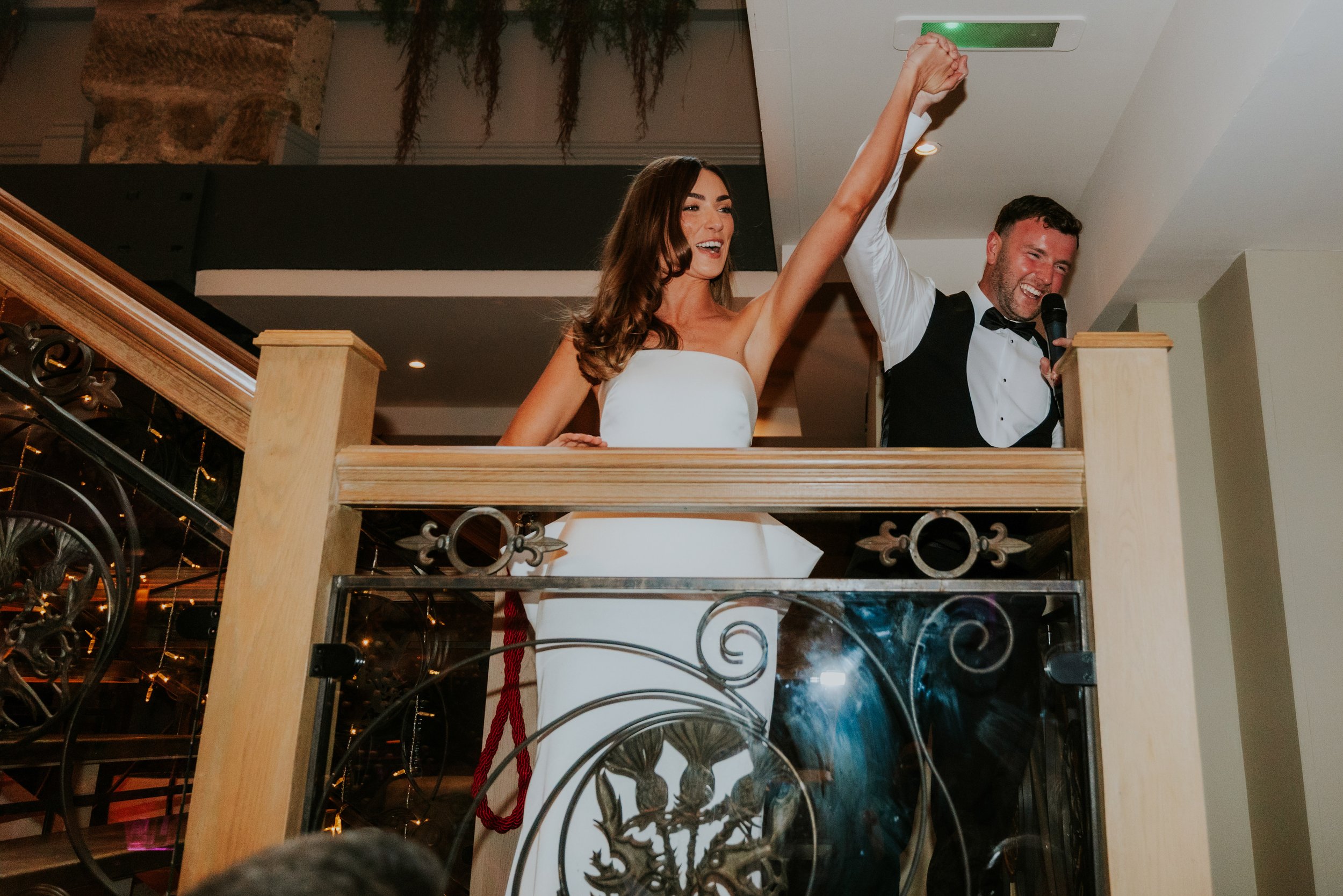 Citation Glasgow wedding photographer 76 Bride and groom on an indoor balcony, holding raised hands; groom smiling with a microphone, bride in a strapless dress.