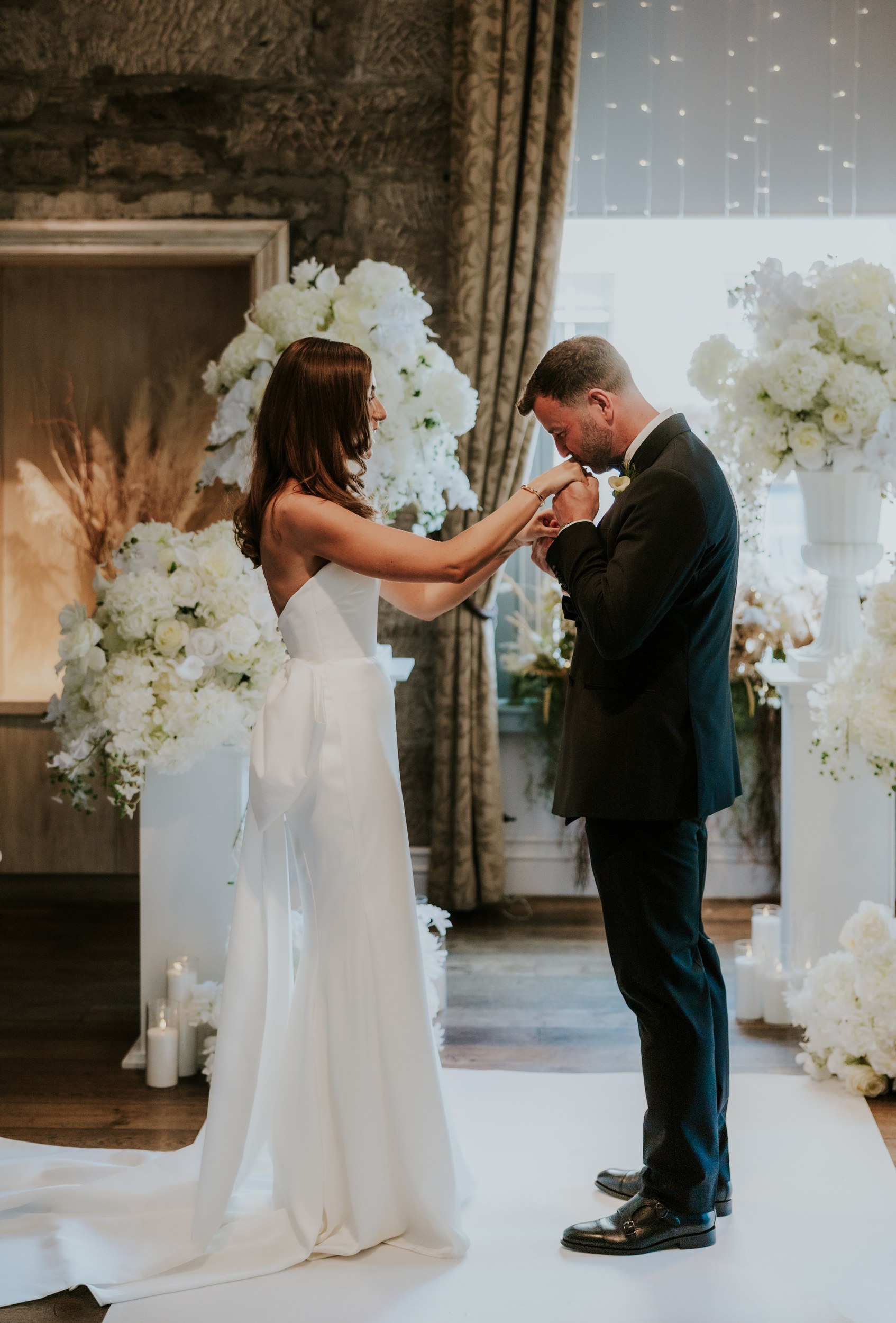 Citation Glasgow wedding photographer 35 Bride and groom share a hand-kissing moment at an indoor ceremony at Citation Glasgow with large white flower arrangements.