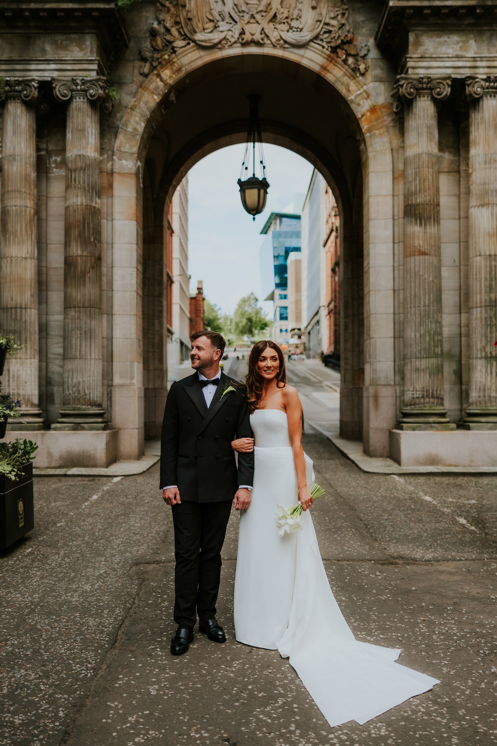 Citation Glasgow wedding photographer 54 Bride and groom under a stone arch at Citation Glasgow; bride in strapless white dress holding lilies, groom in black suit.