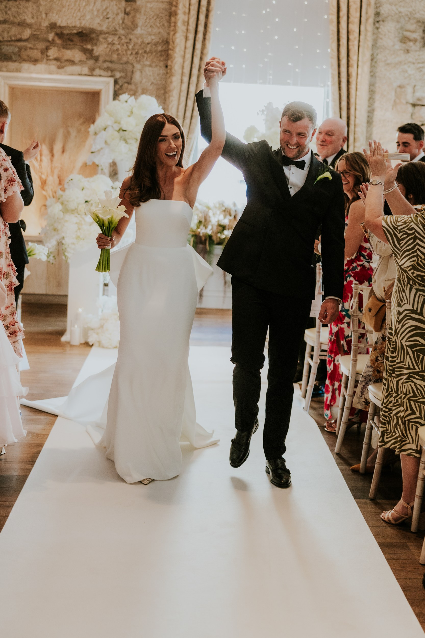 Citation Glasgow wedding photographer 36 Bride and groom walk down the aisle hand-in-hand, bride holding calla lilies as guests clap in a stone-walled ceremony room.
