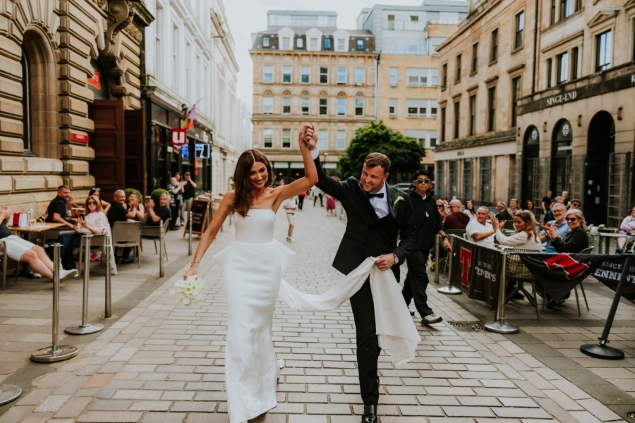 Bride and groom walk hand-in-hand down a Glasgow street outside Citation Glasgow while café guests applaud.