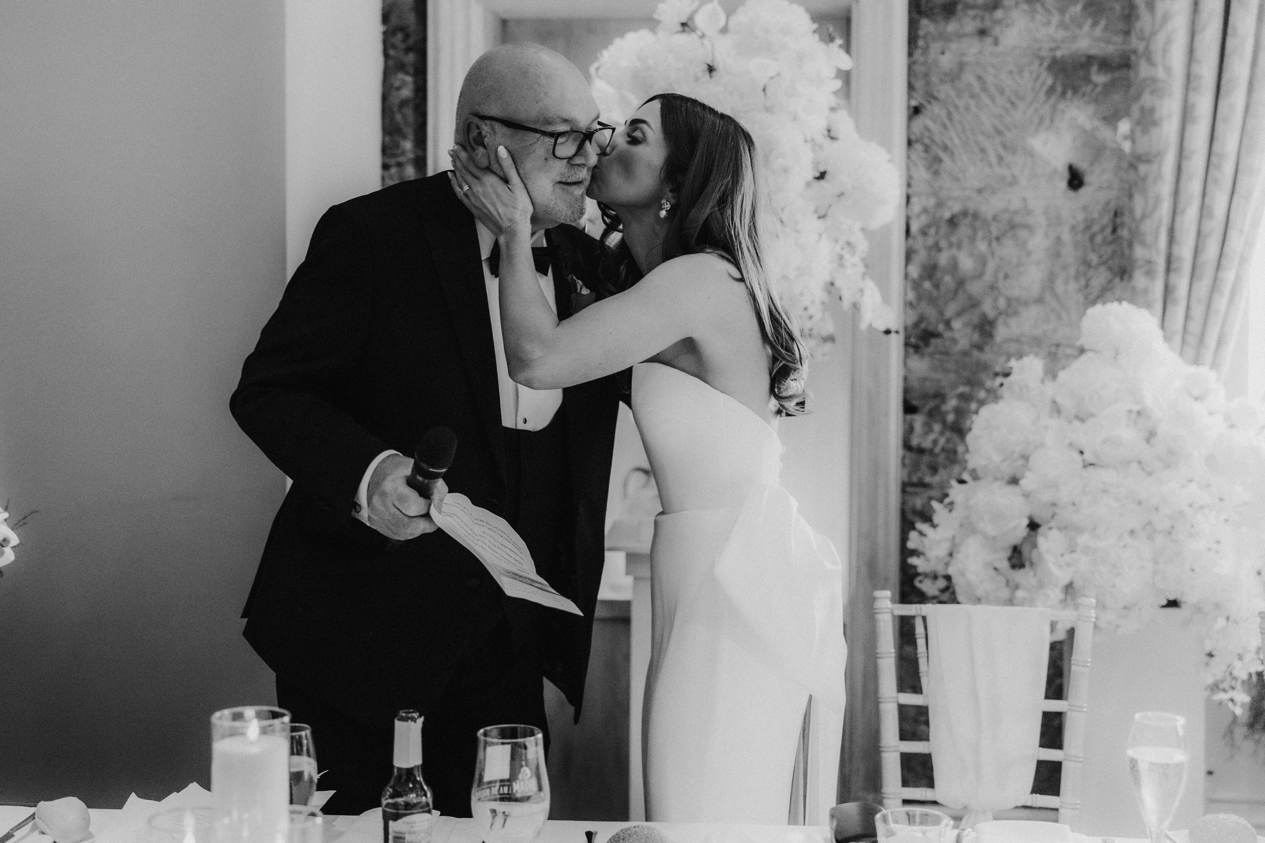 Citation Glasgow wedding photographer 75 Bride Colette kisses a man holding a microphone during a reception speech, with glasses and floral arrangements on the table behind them.