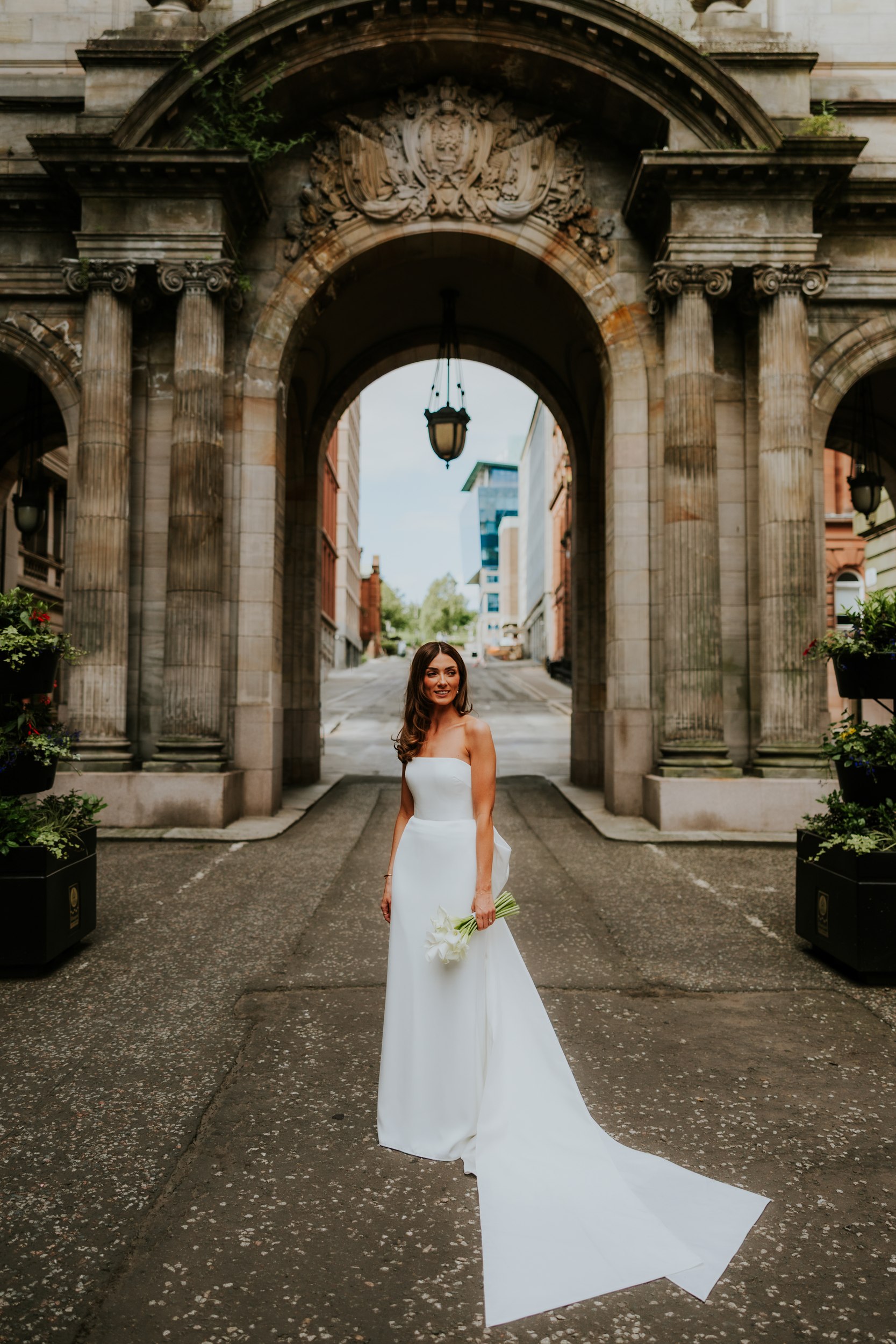 Citation Glasgow wedding photographer 56 Bride Colette Maguire in strapless white gown holding lilies beneath a carved stone arch at Citation Glasgow.