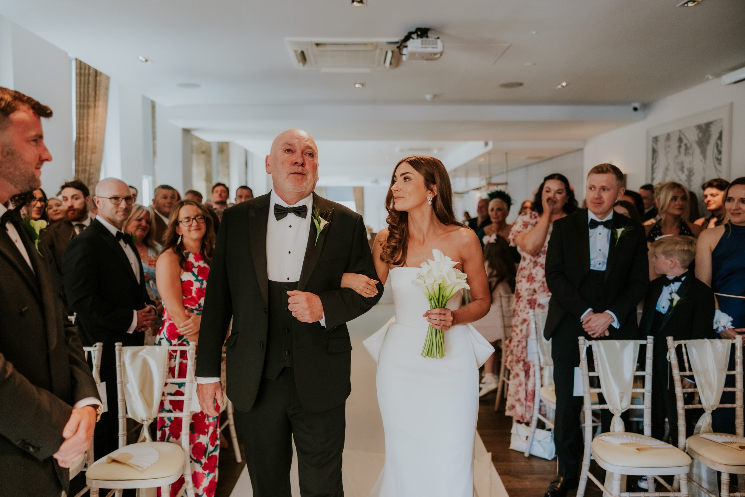 Citation Glasgow wedding photographer 25 Bride holding white calla lilies escorted down the aisle by a man in a tuxedo while guests watch.