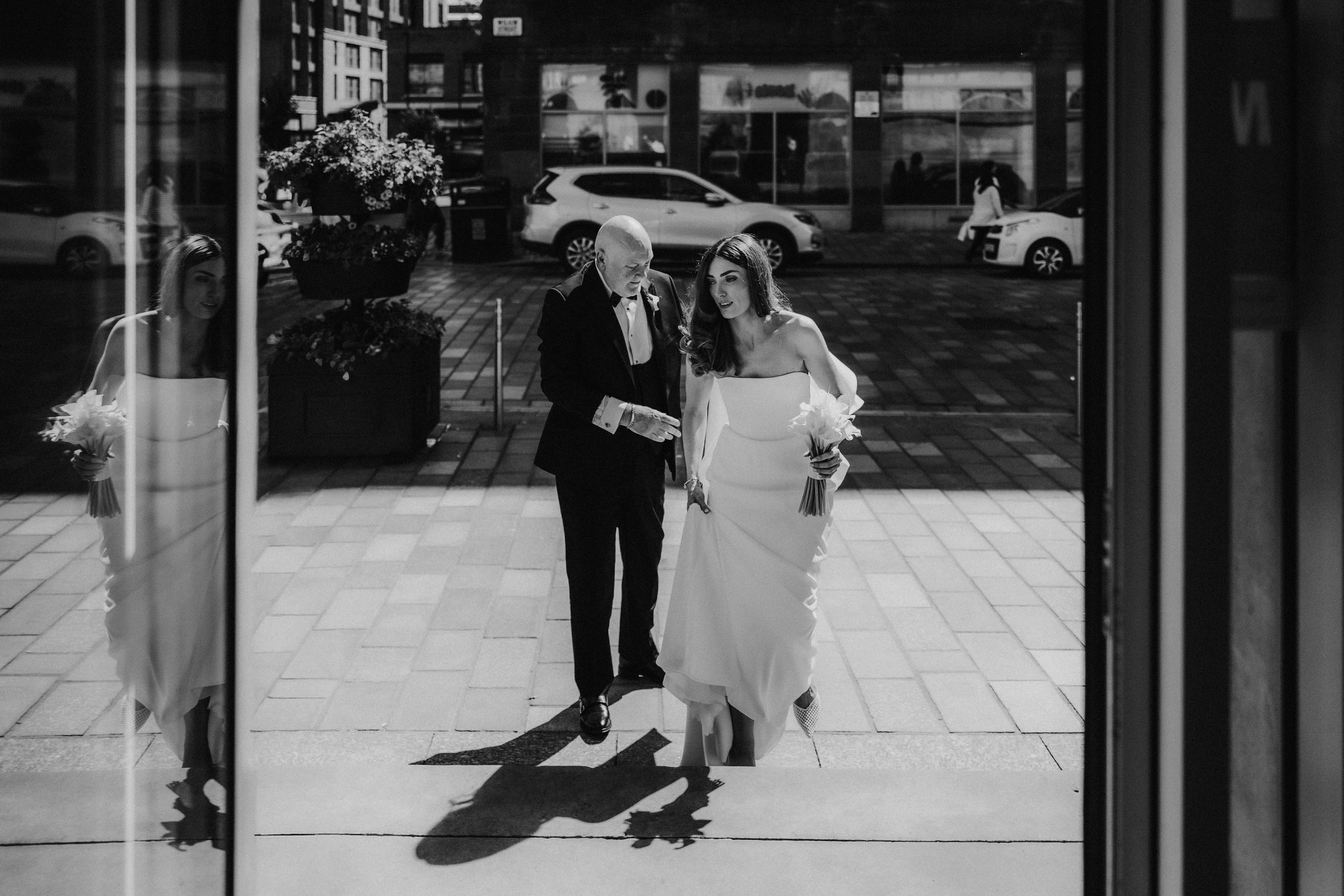 Citation Glasgow wedding photographer 22 Bride in a strapless dress with bouquet, escorted by a man in a tux outside a city venue, with glass reflections and parked cars behind them.