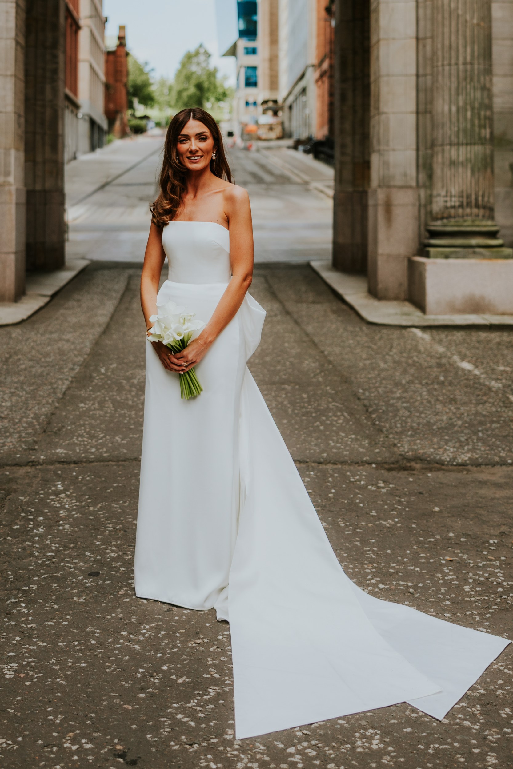 Citation Glasgow wedding photographer 55 Bride in a strapless wedding dress holding calla lilies beneath a stone arch on a city street.