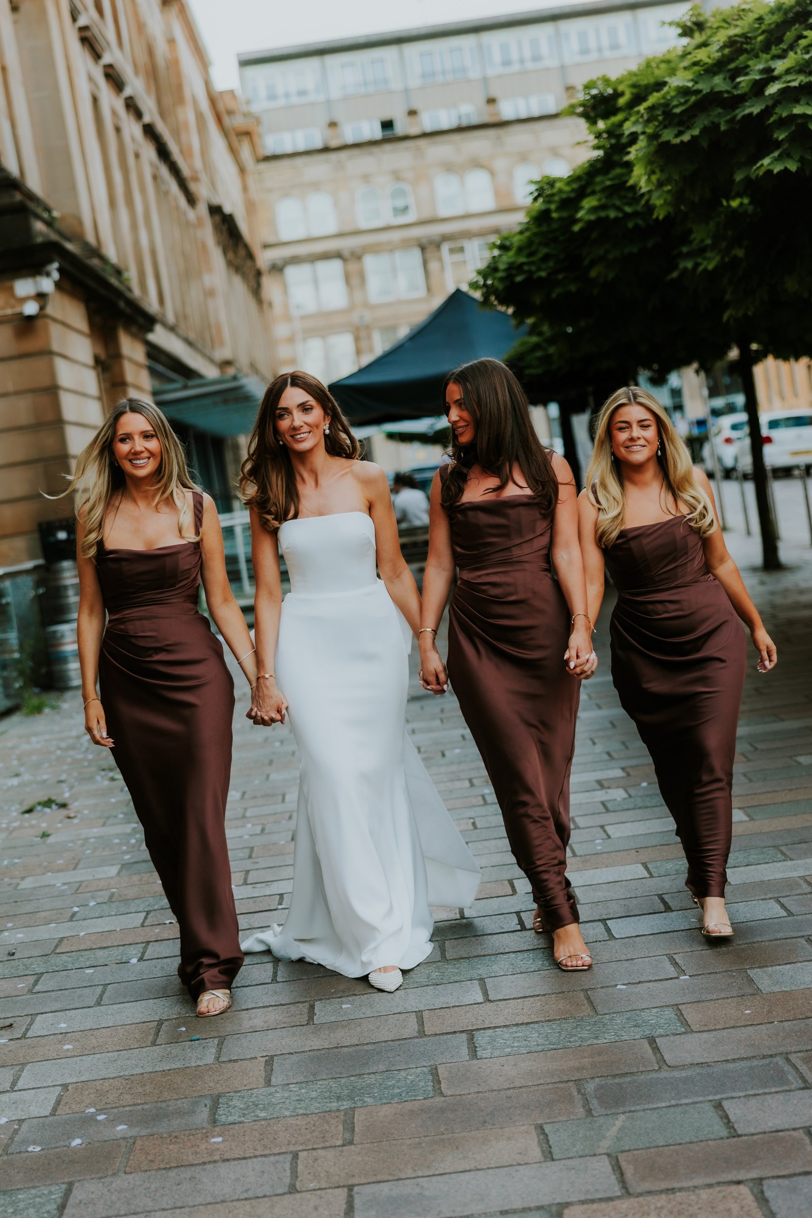 Citation Glasgow wedding photographer 91 Bride in a strapless white gown walking hand-in-hand with three bridesmaids in matching brown dresses on a city pavement.