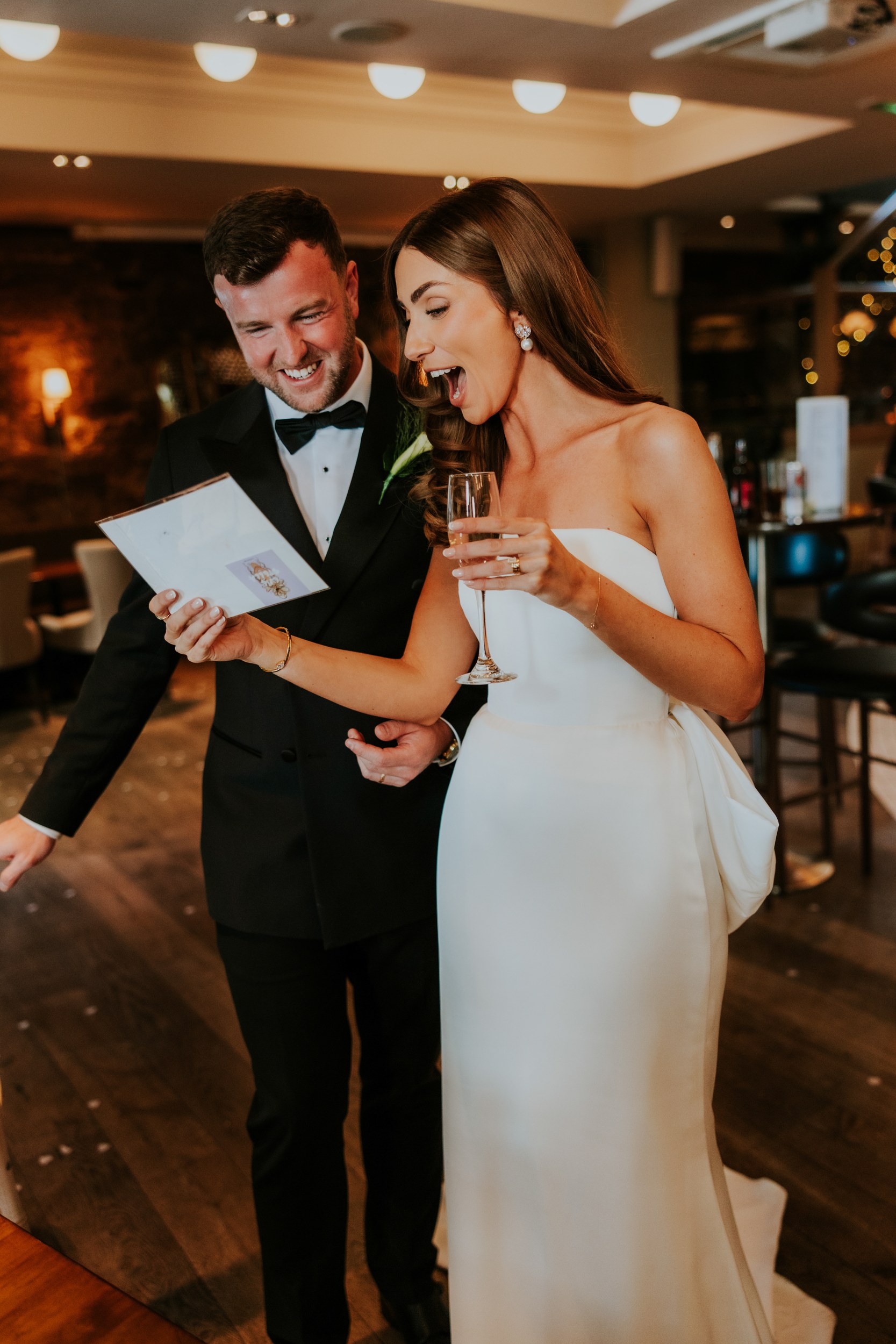 Citation Glasgow wedding photographer 67 Bride in strapless dress and groom in tux read a card and laugh, bride holding a champagne glass at their reception.