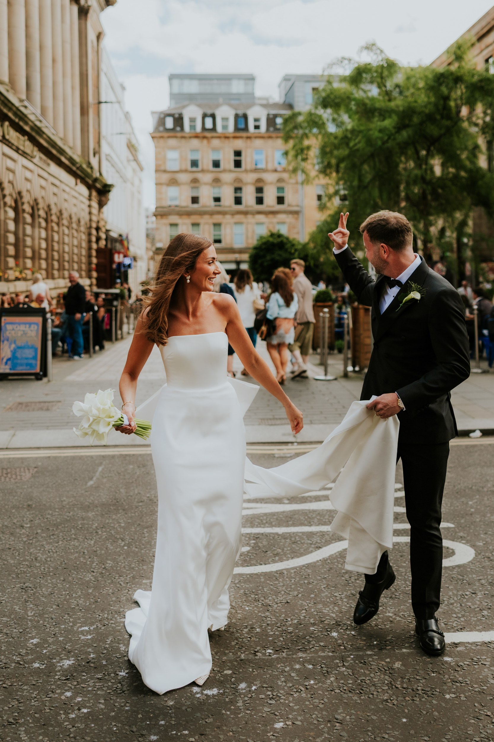 Citation Glasgow wedding photographer 49 Bride in strapless gown with white calla lilies, groom in tux lifts her train as they cross a Glasgow street outside Citation Glasgow.