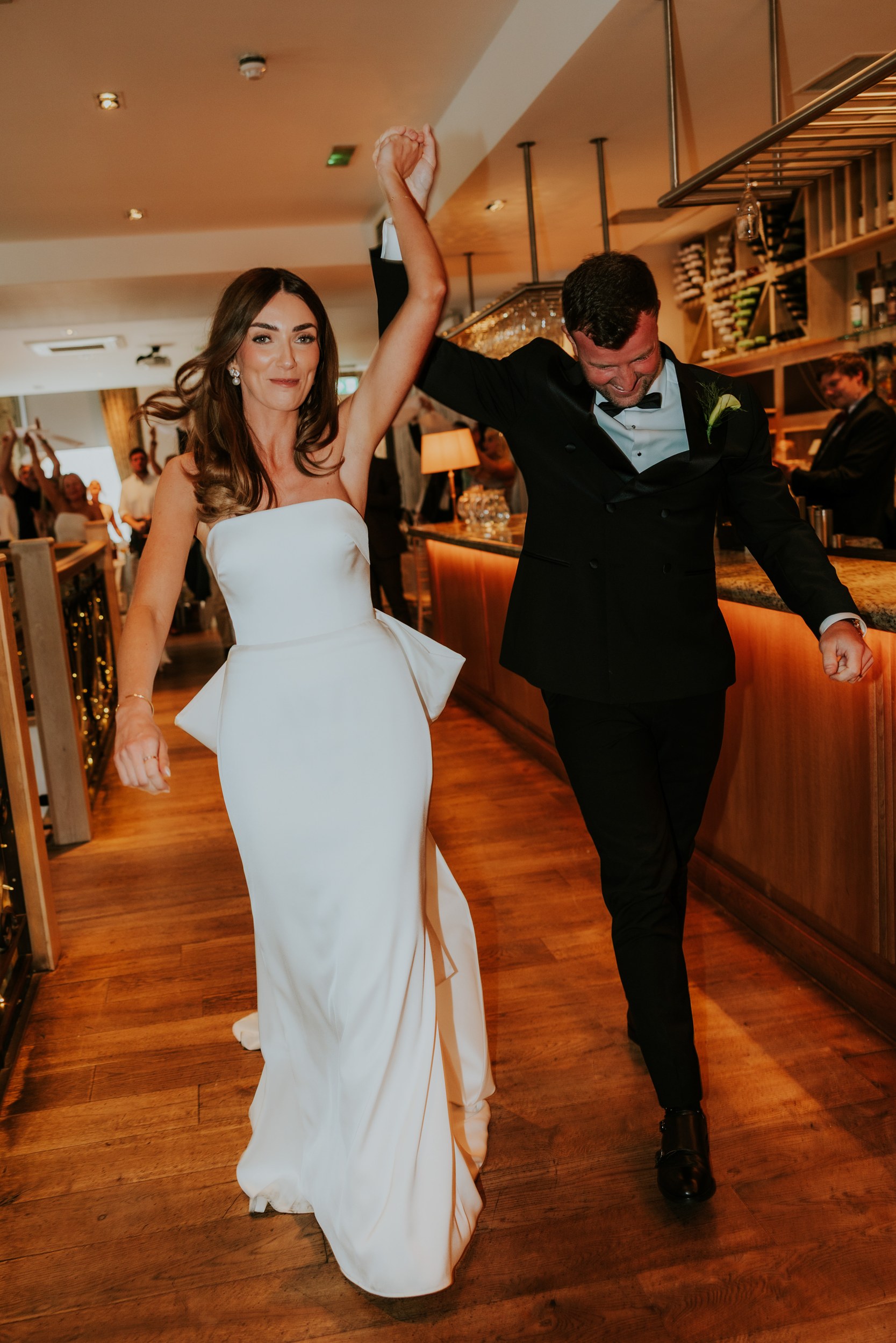 Citation Glasgow wedding photographer 72 Bride in strapless white dress and groom in black tux raise joined hands while entering a warm-lit bar area as guests cheer.
